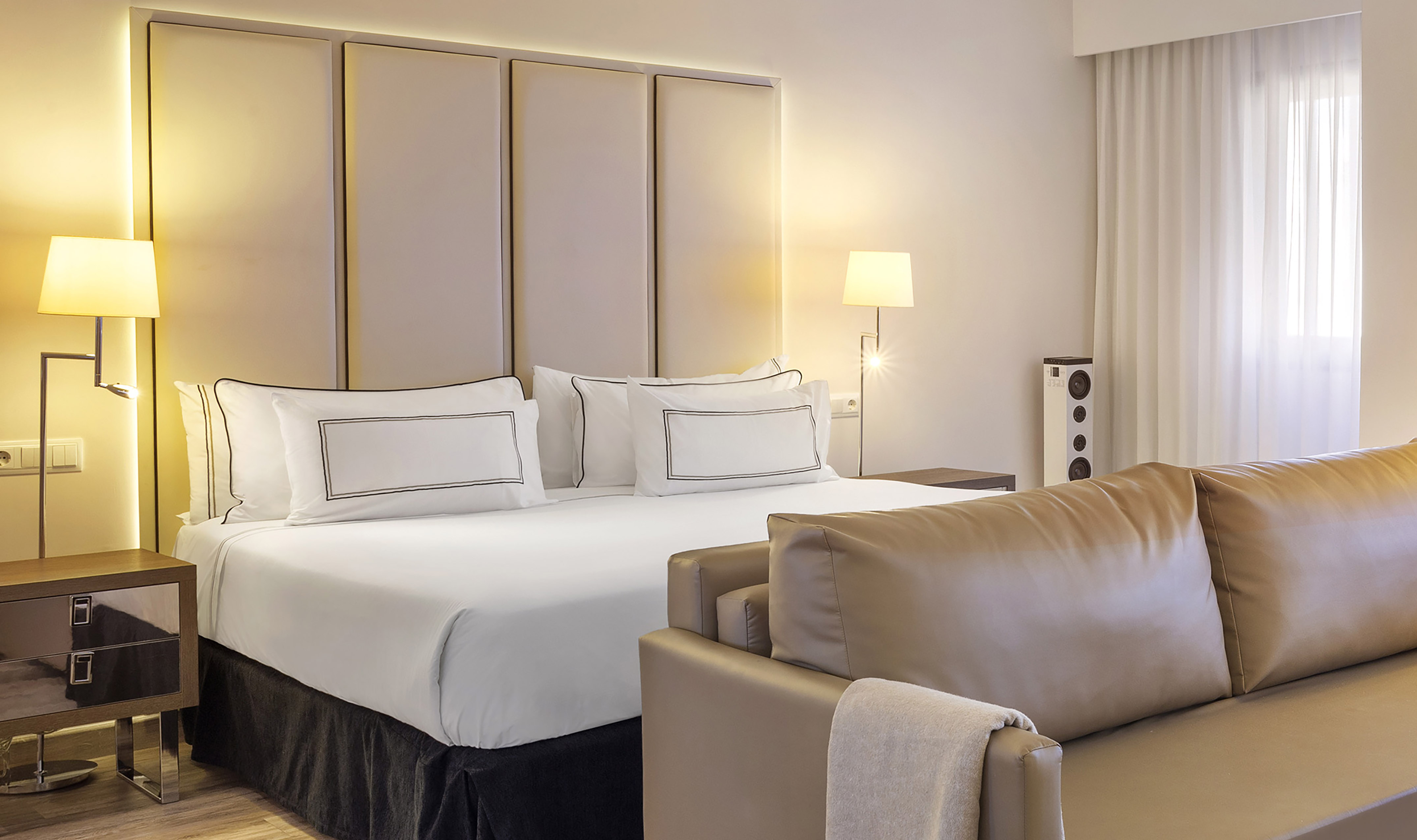 Modern hotel room with large bed, white linens, padded headboard, and beige sofa.