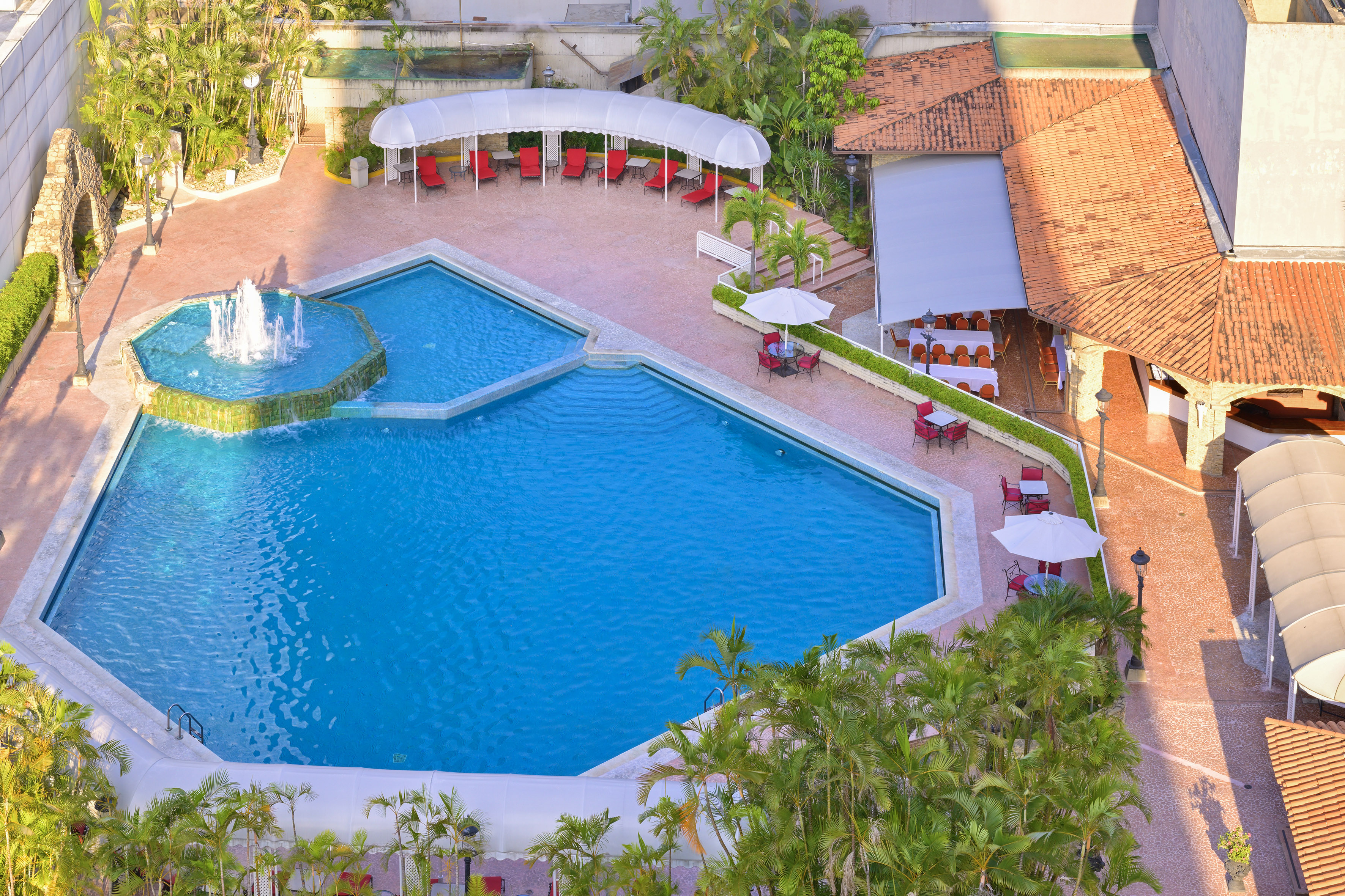 Resort swimming pool with fountain, lounge chairs, palms, and outdoor cafe.