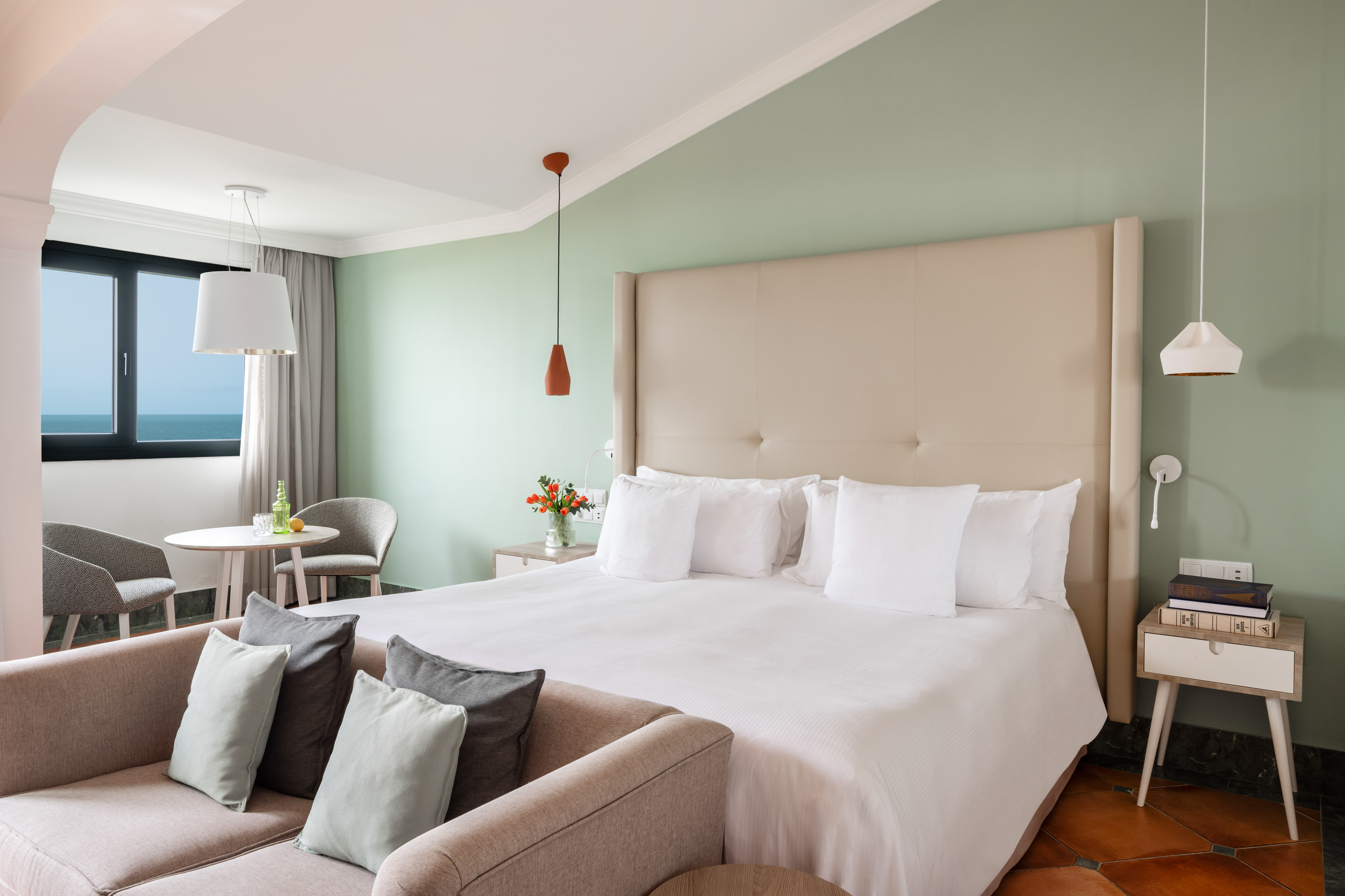 Modern hotel bedroom with king bed, sofa, and sea view from window.