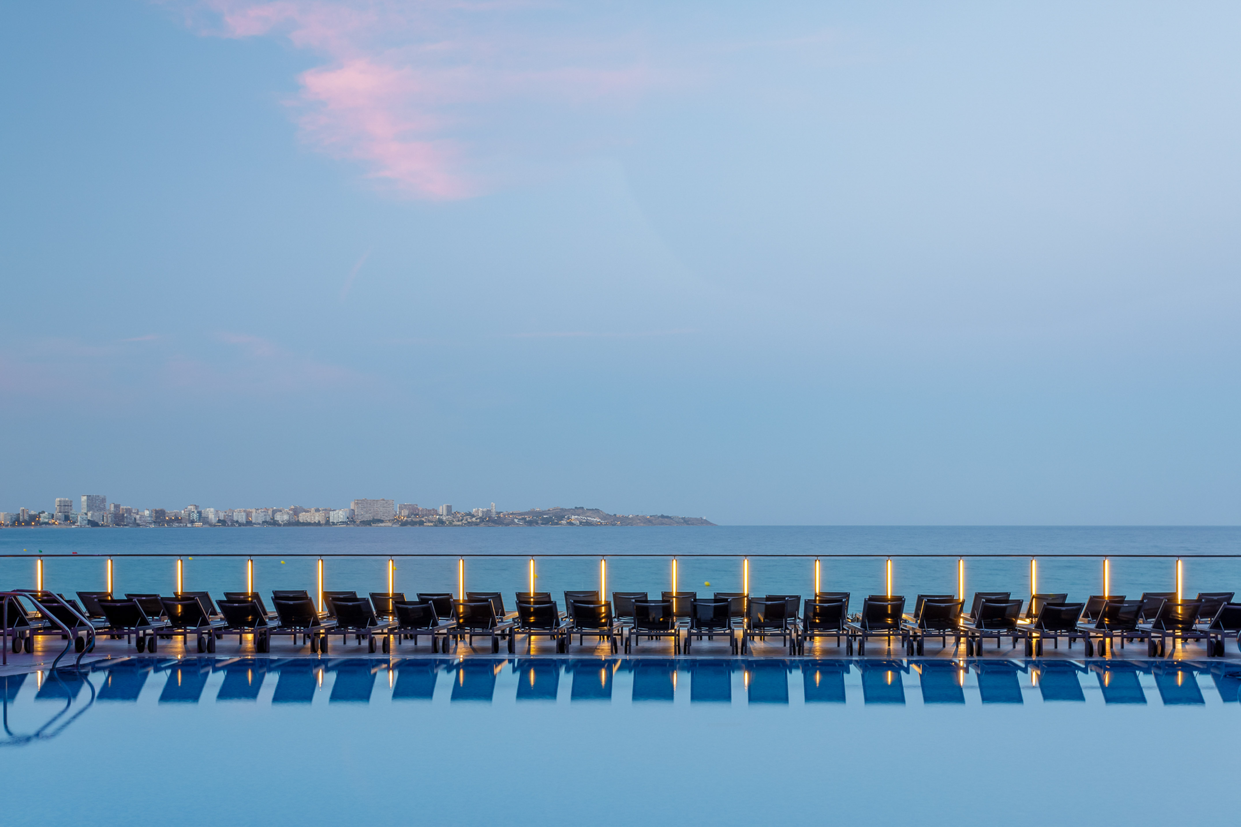 Dusk view of infinity pool, illuminated lounge chairs, ocean, and city.