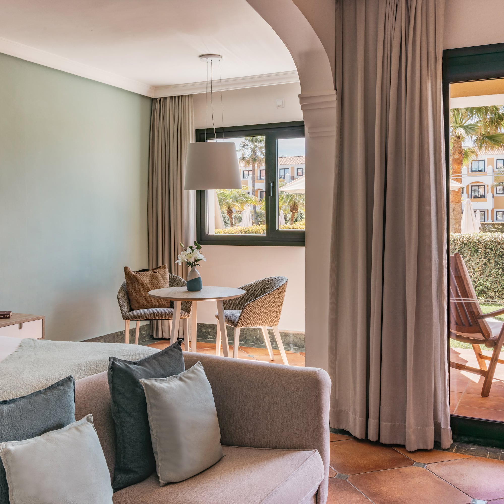 Bright hotel room with bed, sofa, dining area, and resort garden patio.