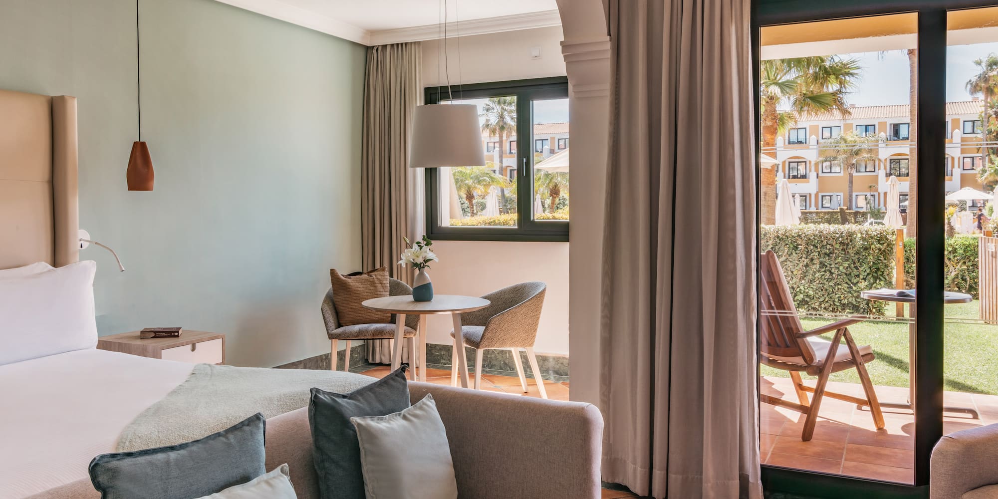 Bright hotel room with bed, sofa, dining area, and resort garden patio.