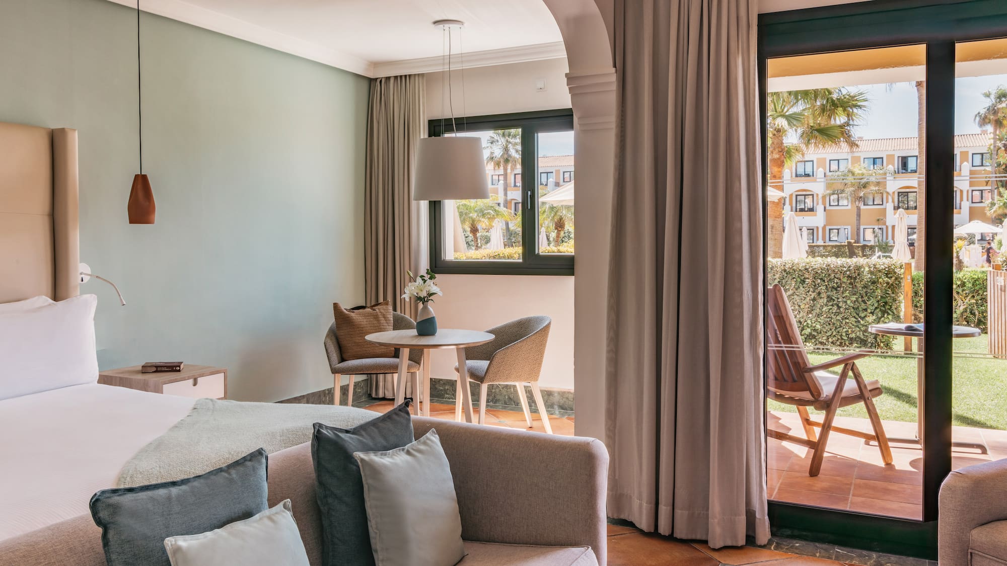 Bright hotel room with bed, sofa, dining area, and resort garden patio.