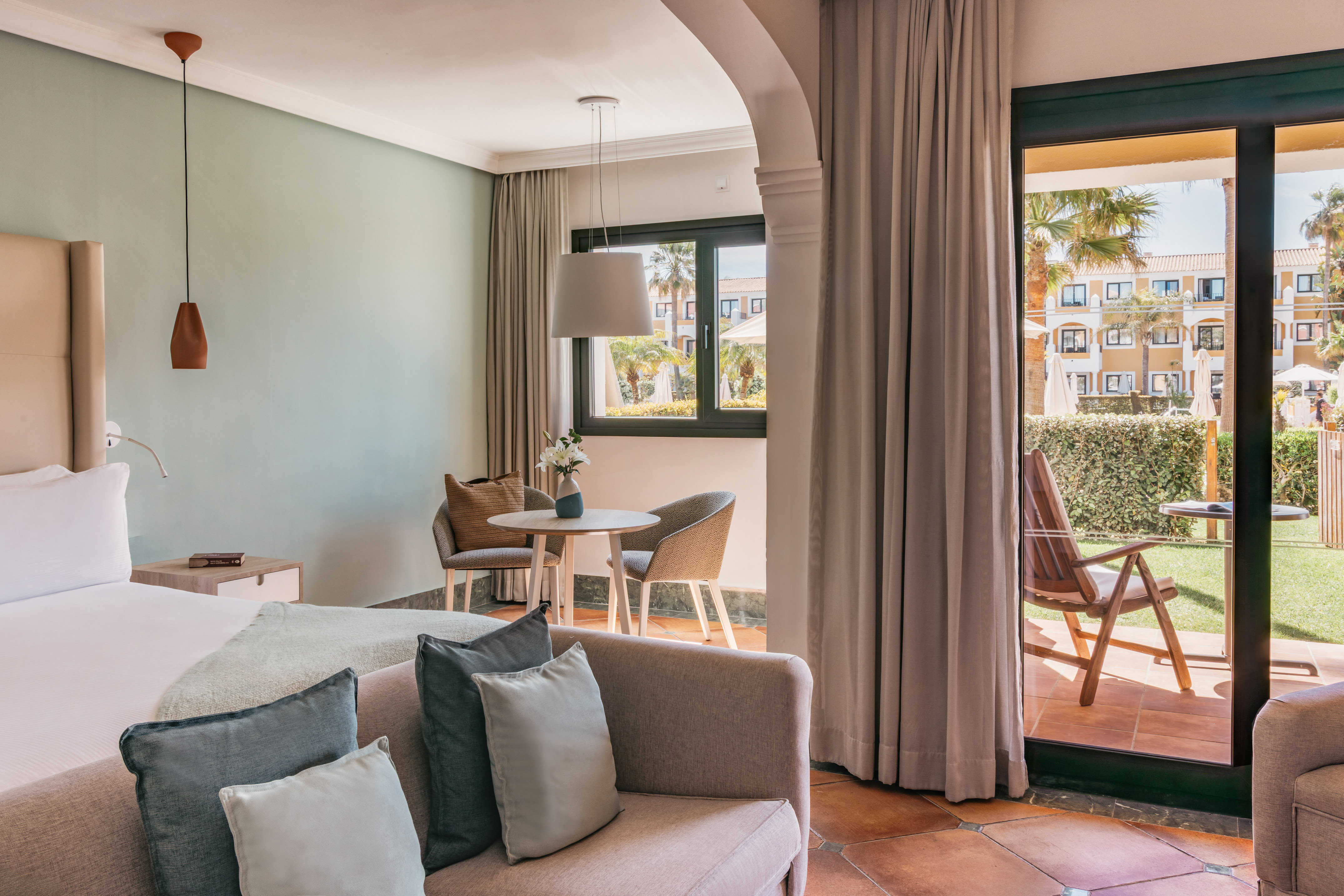 Bright hotel room with bed, sofa, dining area, and resort garden patio.