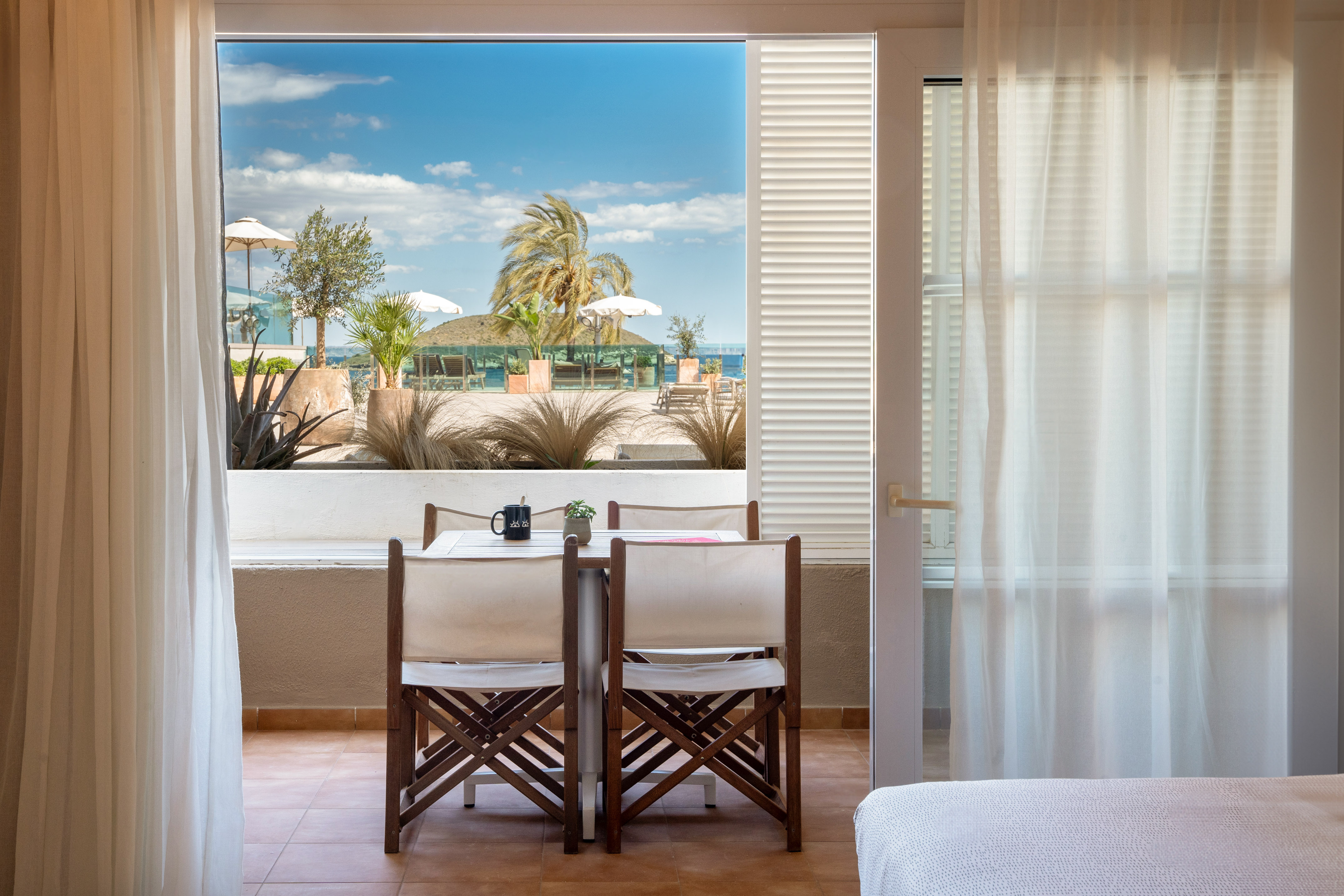 Hotel room balcony with table and chairs, overlooking a sunny beach and palm trees.