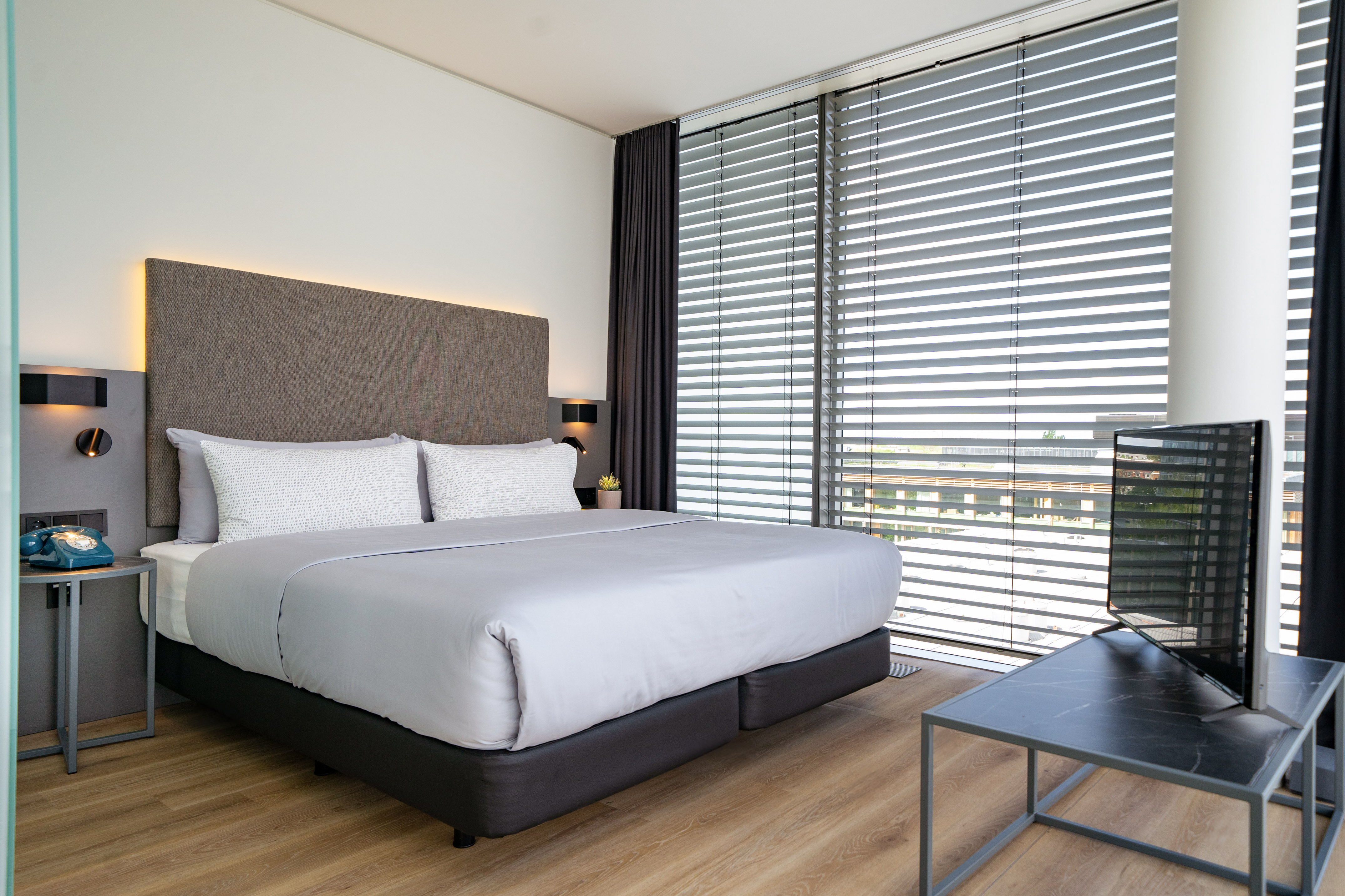Modern hotel room with large grey bed, backlit headboard, and window blinds.