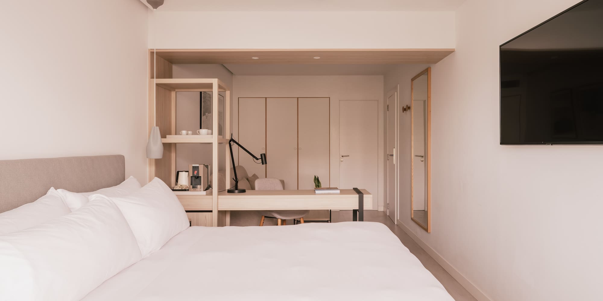 Modern hotel bedroom with white bed, desk, TV, and coffee station.