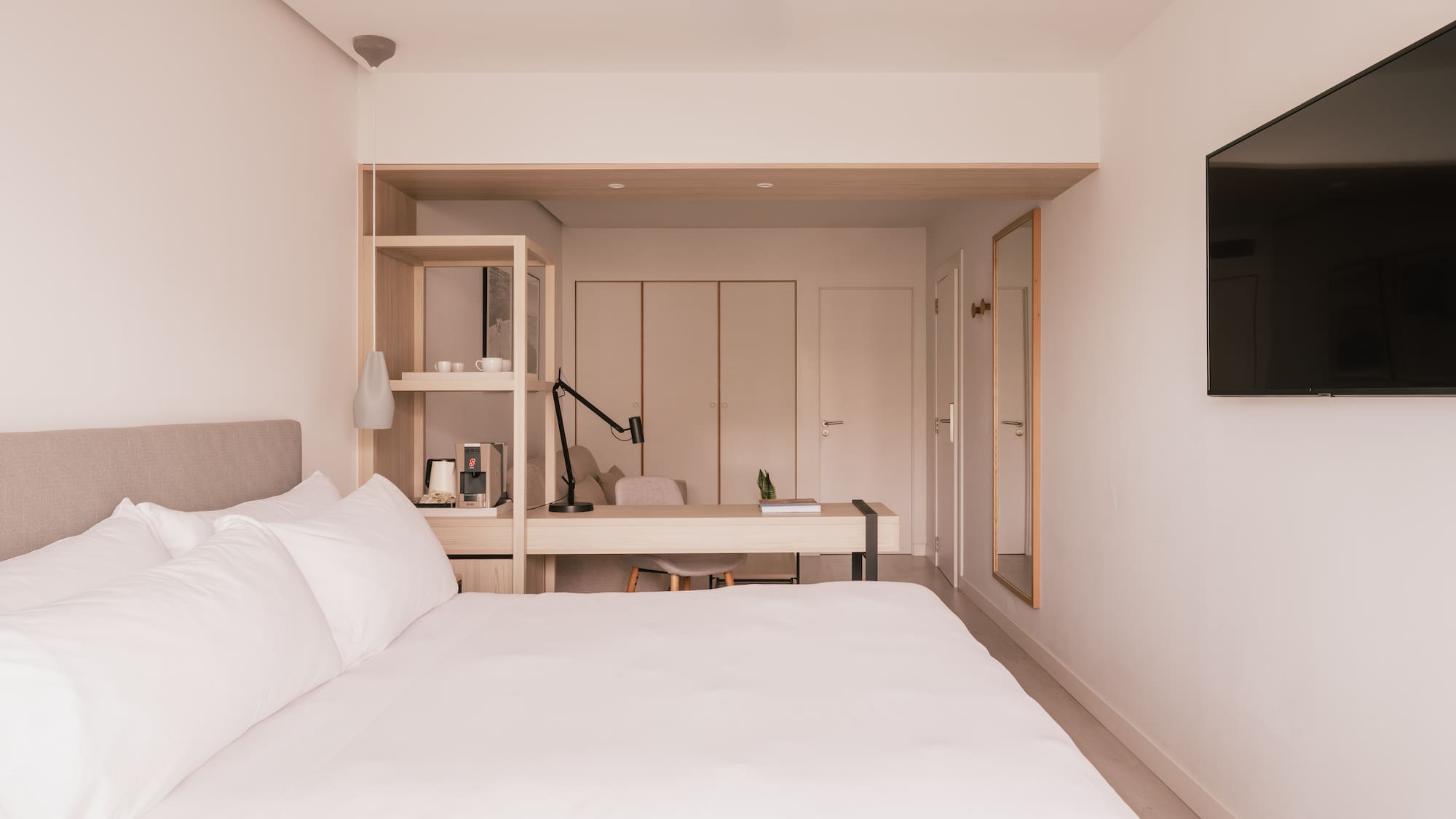 Modern hotel bedroom with white bed, desk, TV, and coffee station.