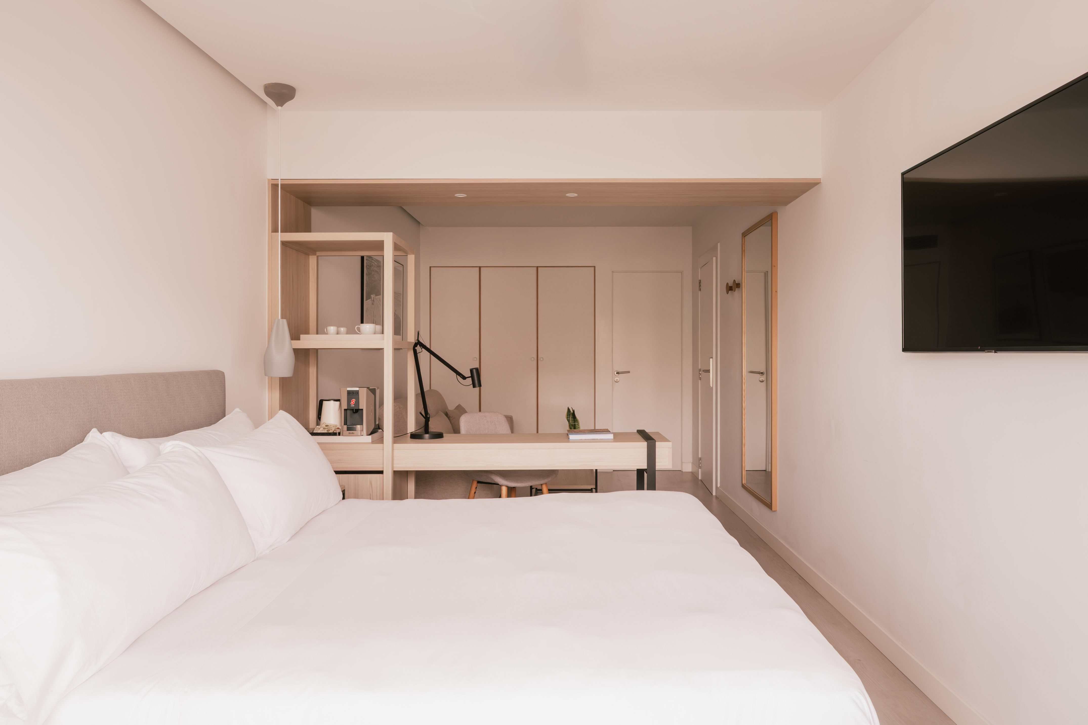 Modern hotel bedroom with white bed, desk, TV, and coffee station.