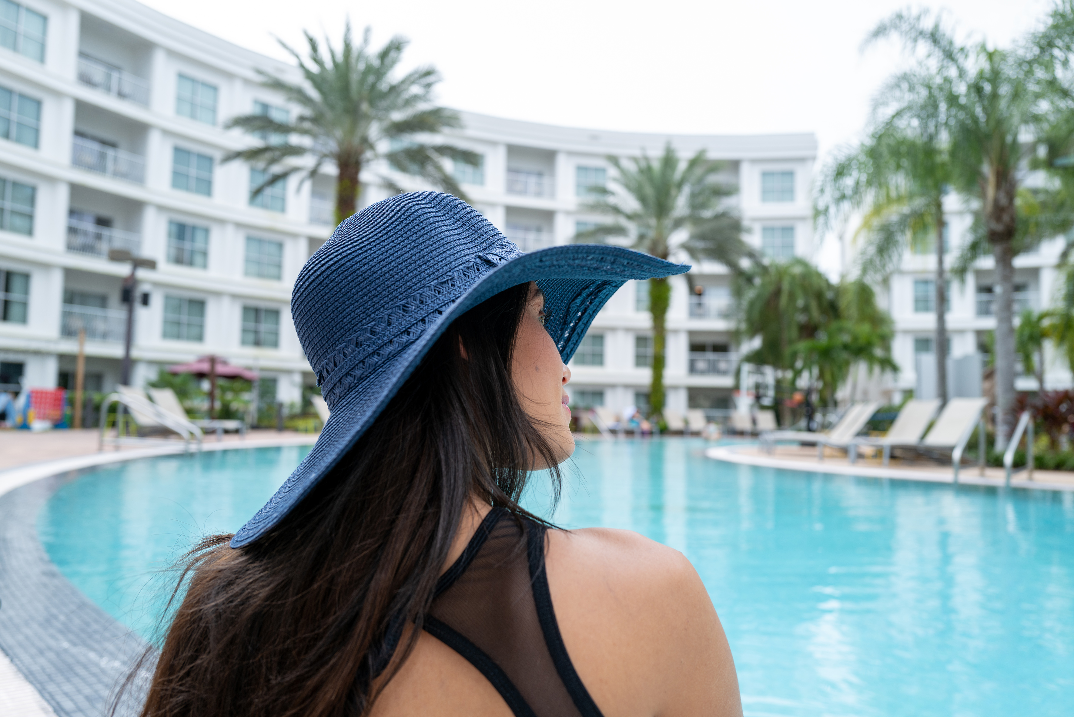 Woman in blue sun hat by resort swimming pool with palm trees.
