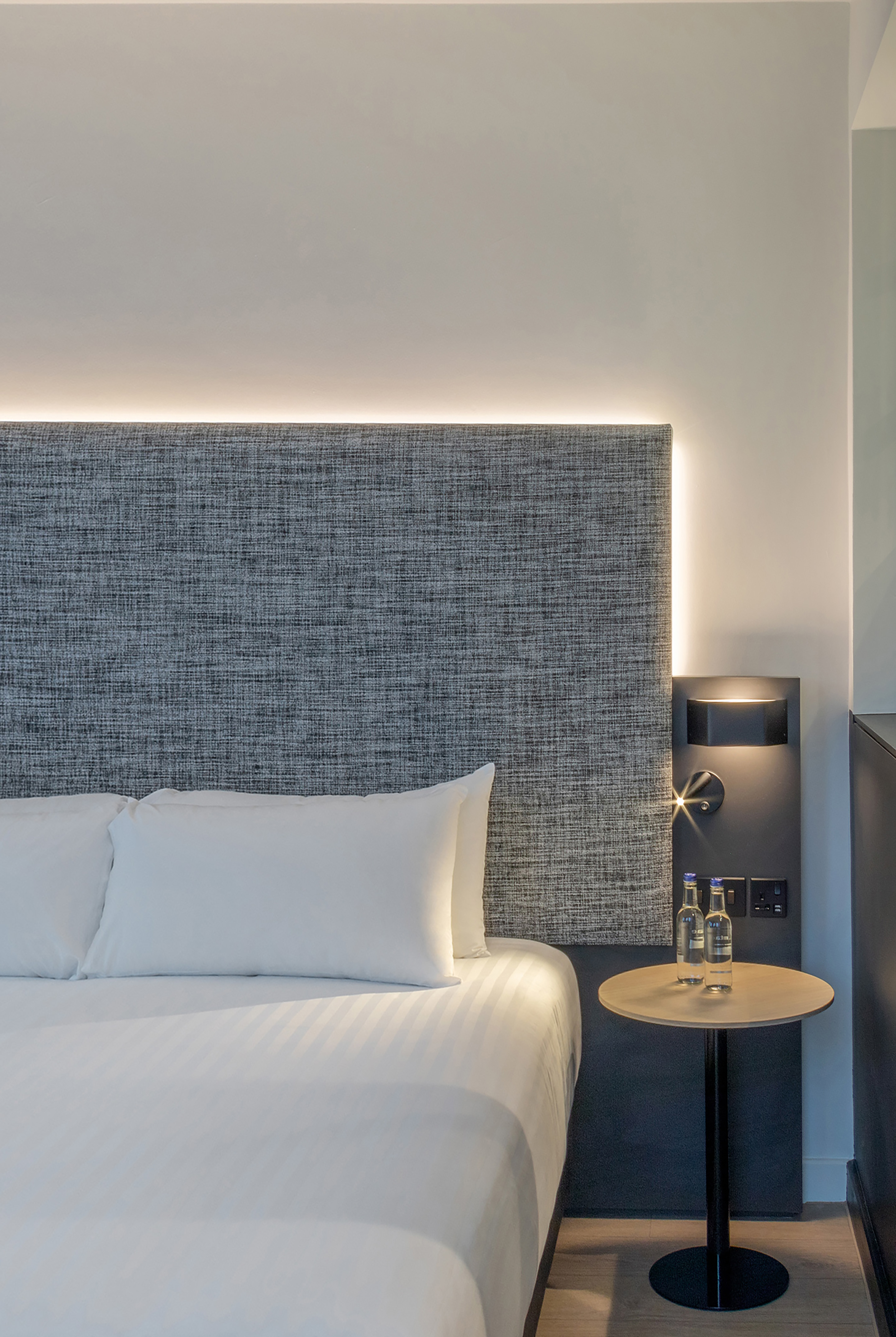 Modern hotel bedroom: white bed, grey headboard with LED light, nightstand.