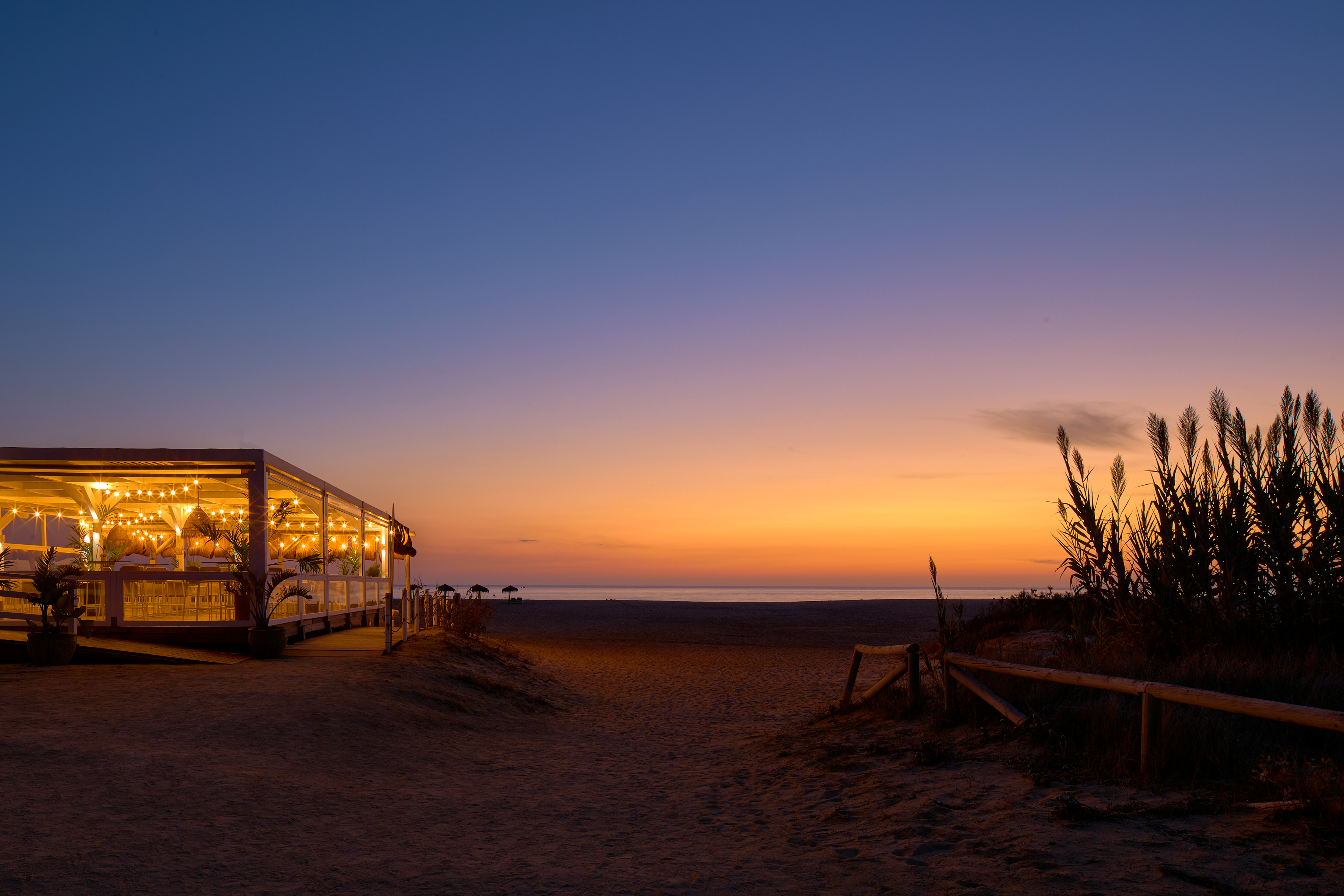 Illuminated beach bar at dusk, sandy path to ocean, colorful sunset sky.
