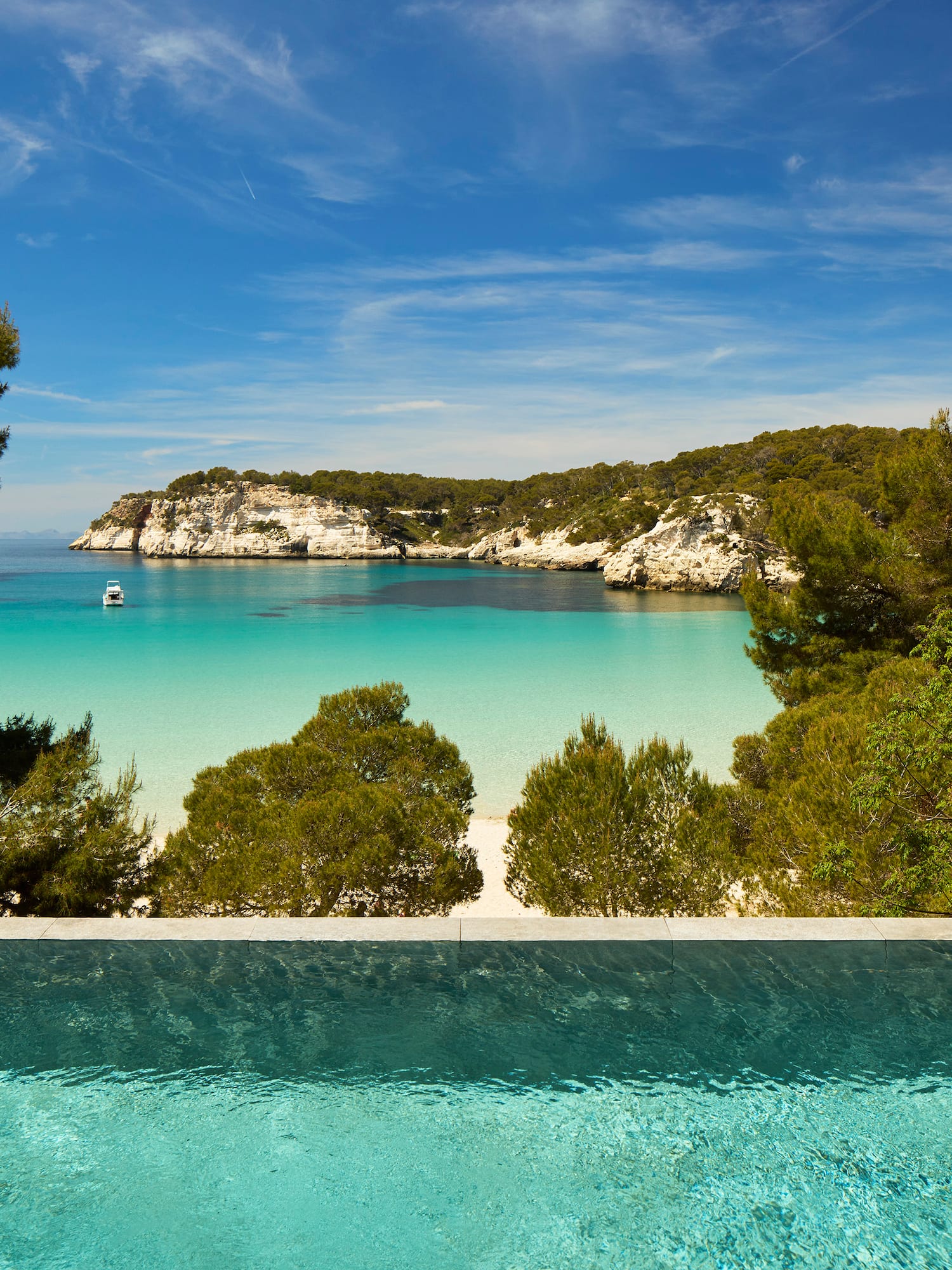 Infinity pool overlooks clear turquoise bay with boat, pine trees, and white cliffs.