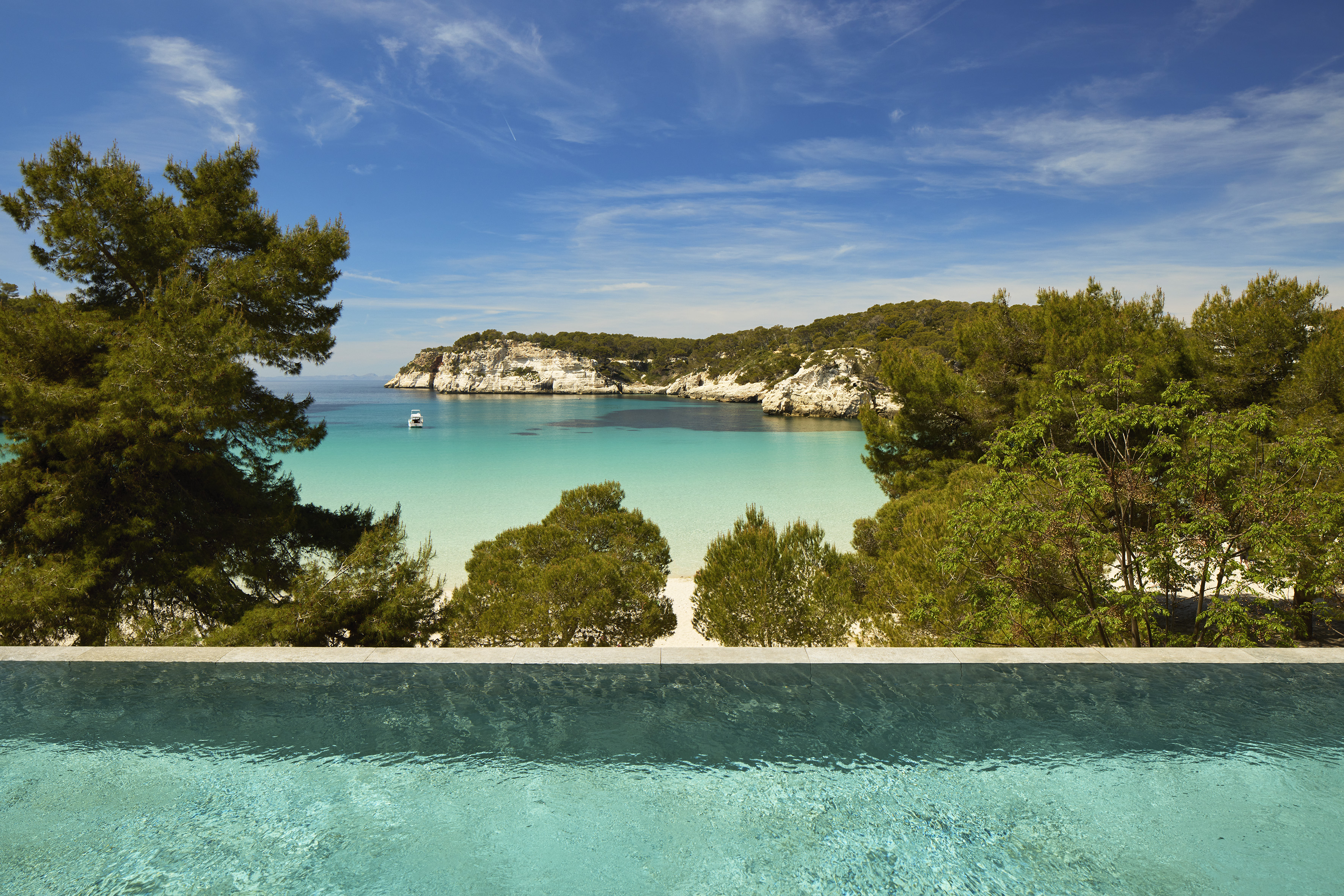 Infinity pool overlooks clear turquoise bay with boat, pine trees, and white cliffs.