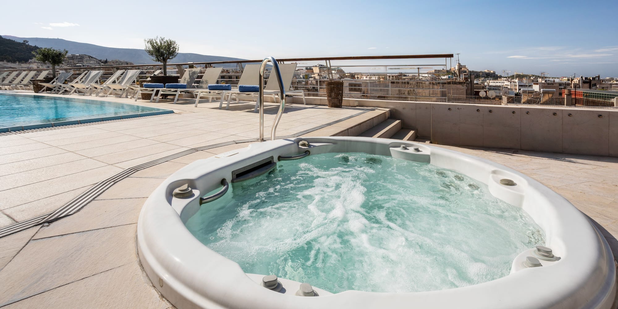 Bubbling rooftop jacuzzi next to swimming pool, sun loungers, and city view.