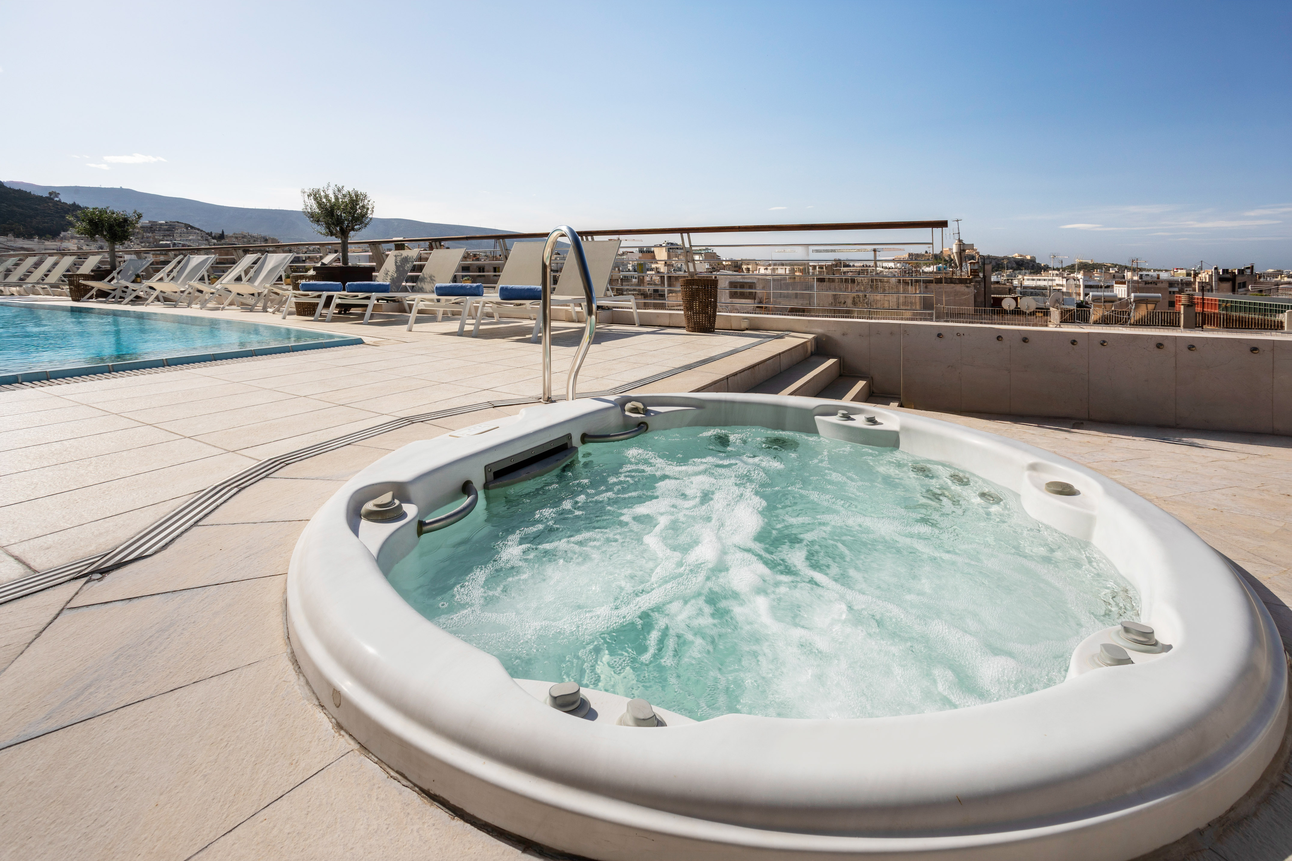Bubbling rooftop jacuzzi next to swimming pool, sun loungers, and city view.