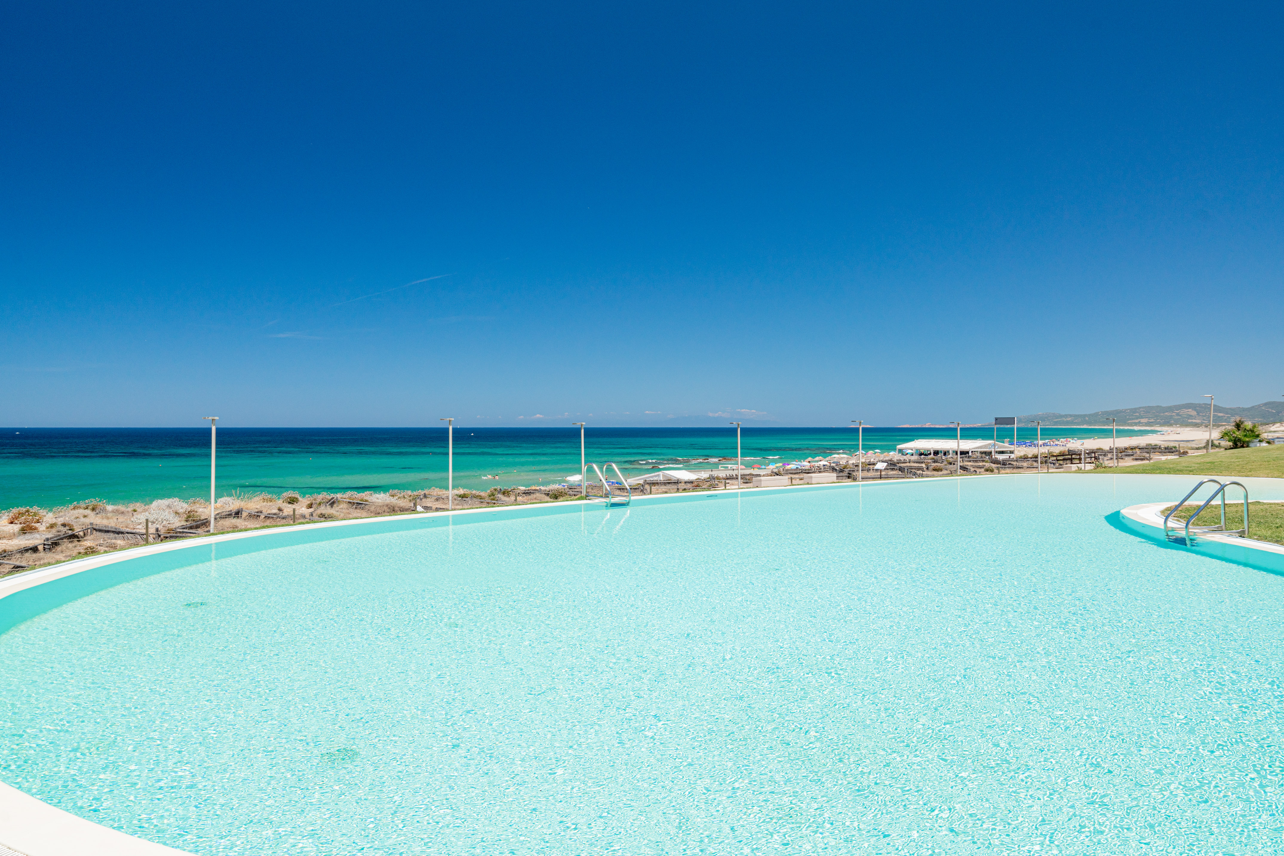 Luxury resort pool with ocean view, sandy beach, and clear blue sky.
