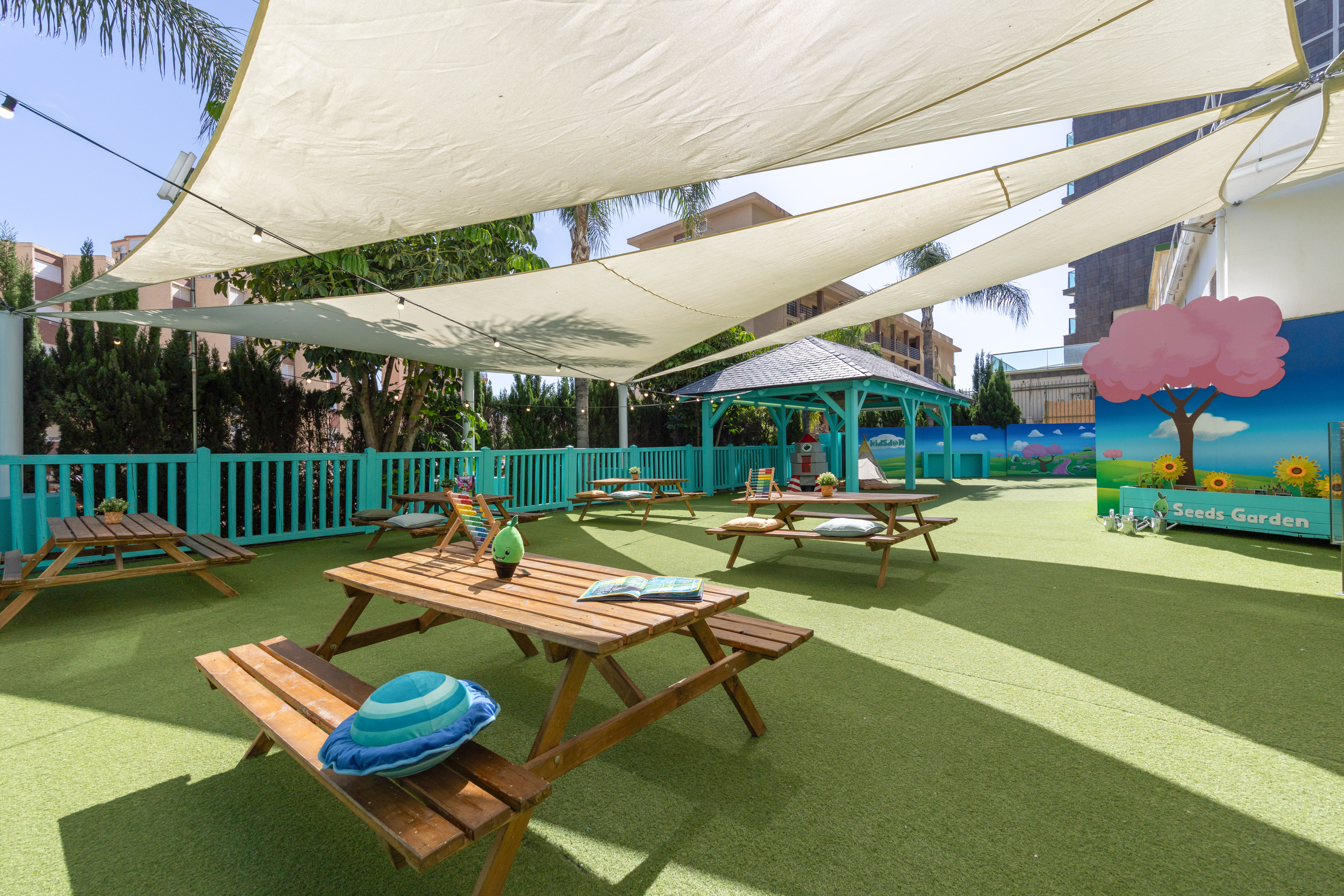 Outdoor kids play area with shade sails, picnic tables, and a 'Seeds Garden' mural.