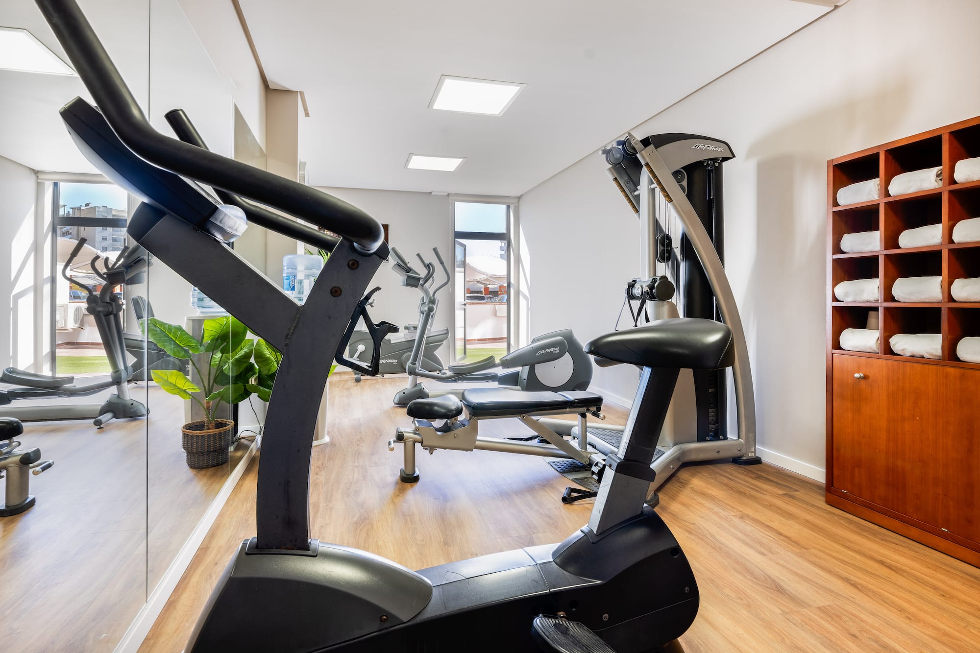 Bright hotel gym with elliptical, stationary bike, weight machine, and towel cabinet.