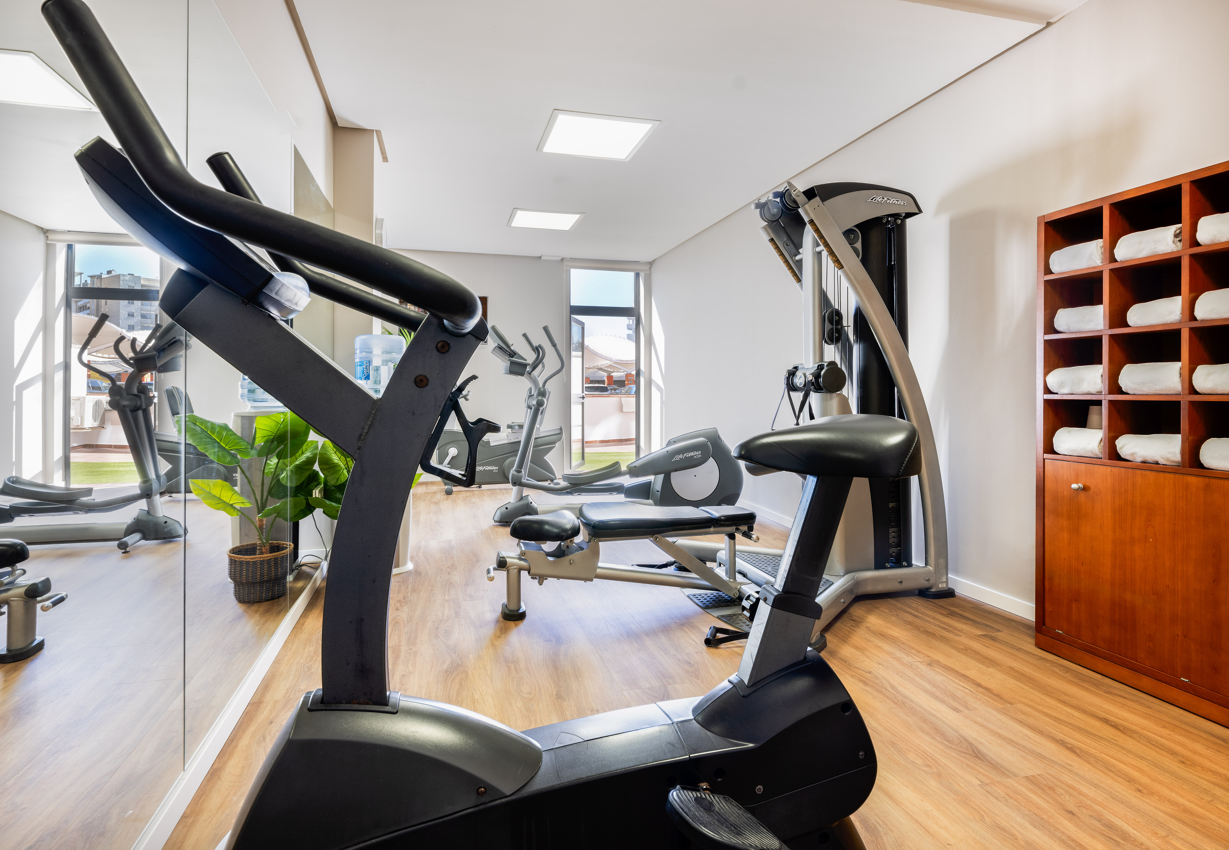 Bright hotel gym with elliptical, stationary bike, weight machine, and towel cabinet.