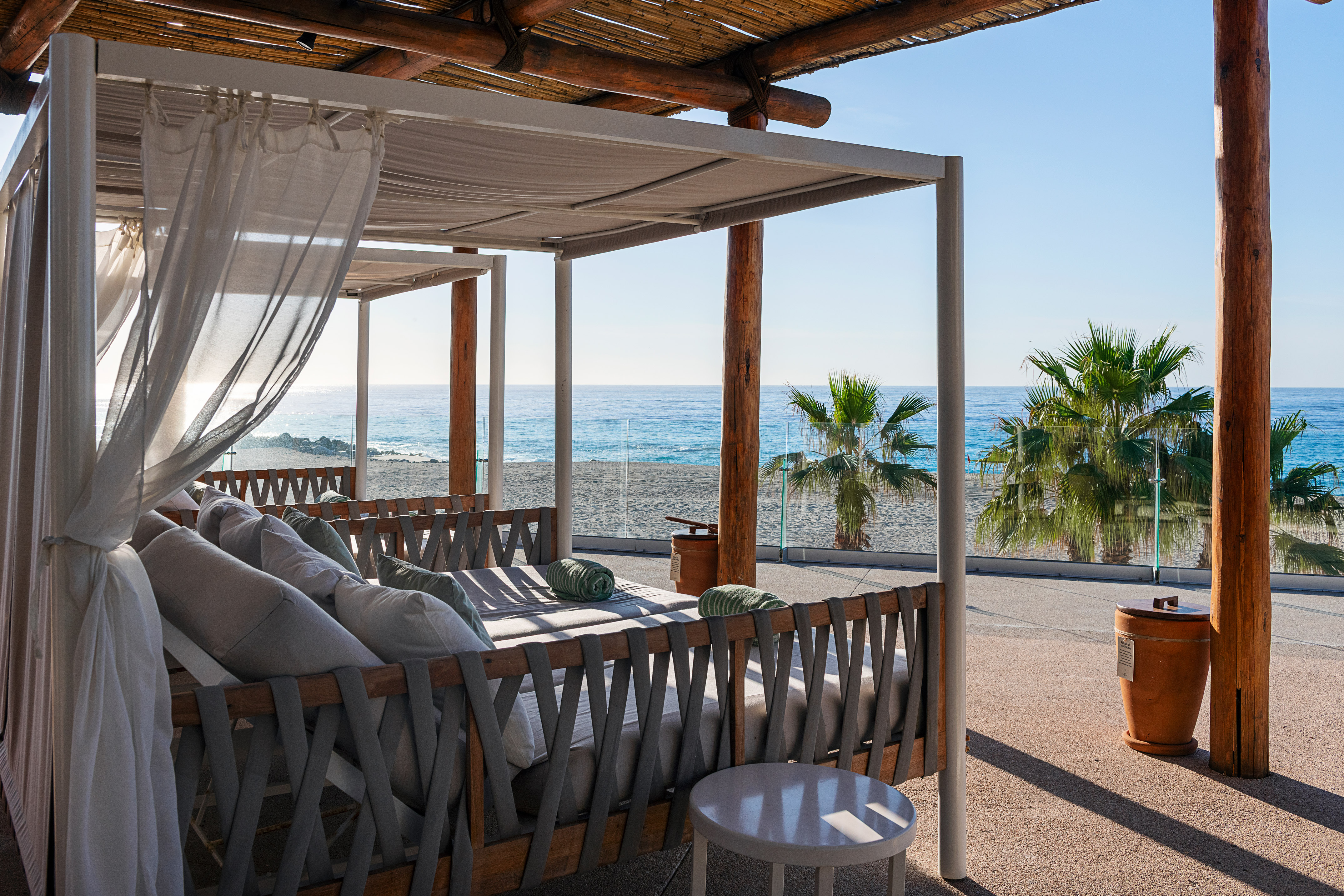 Beachfront daybed cabana with white curtains, ocean, sand, and palm trees.