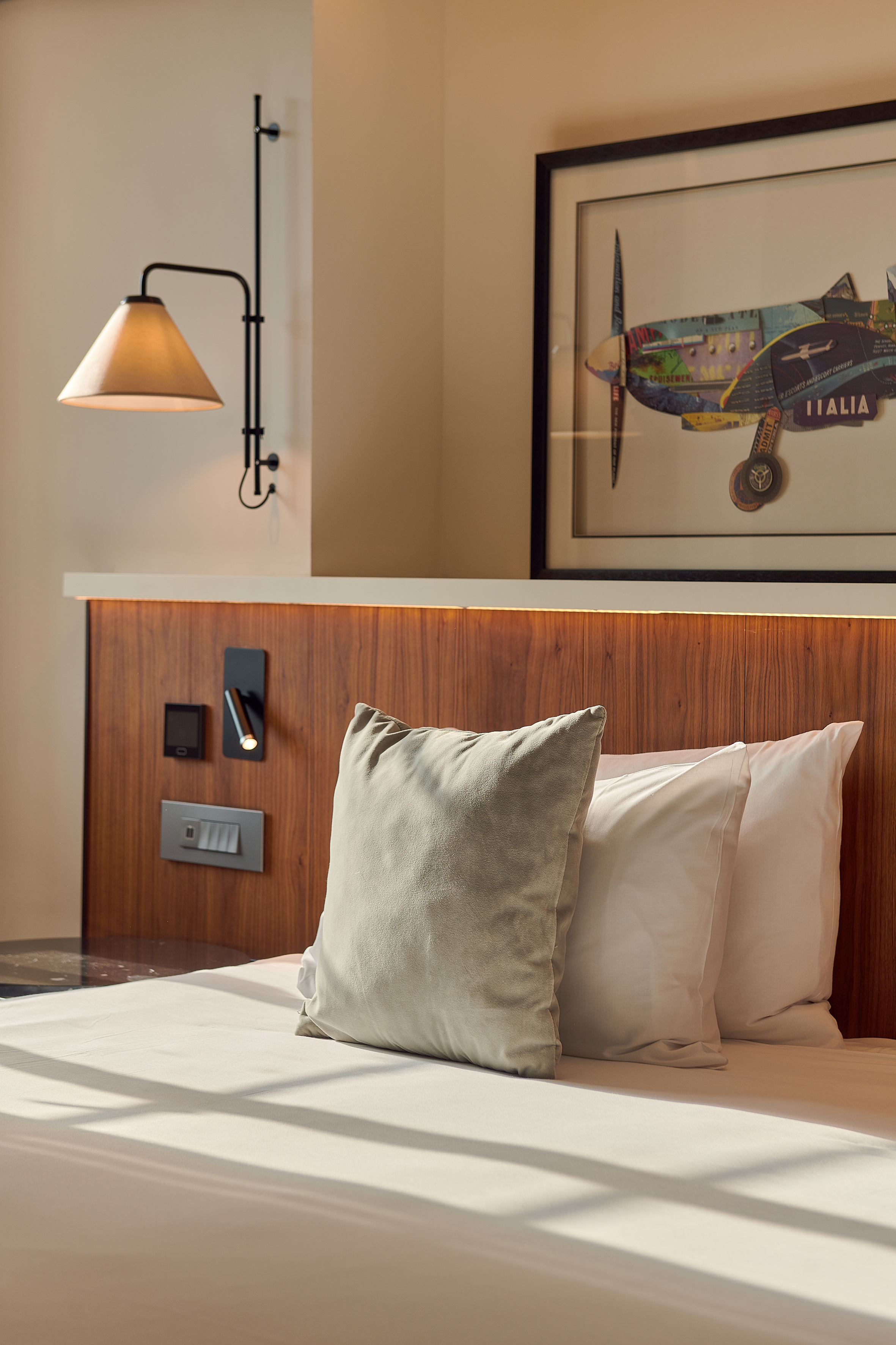 Hotel bed with grey pillow, wooden headboard, wall lamp, and airplane art.