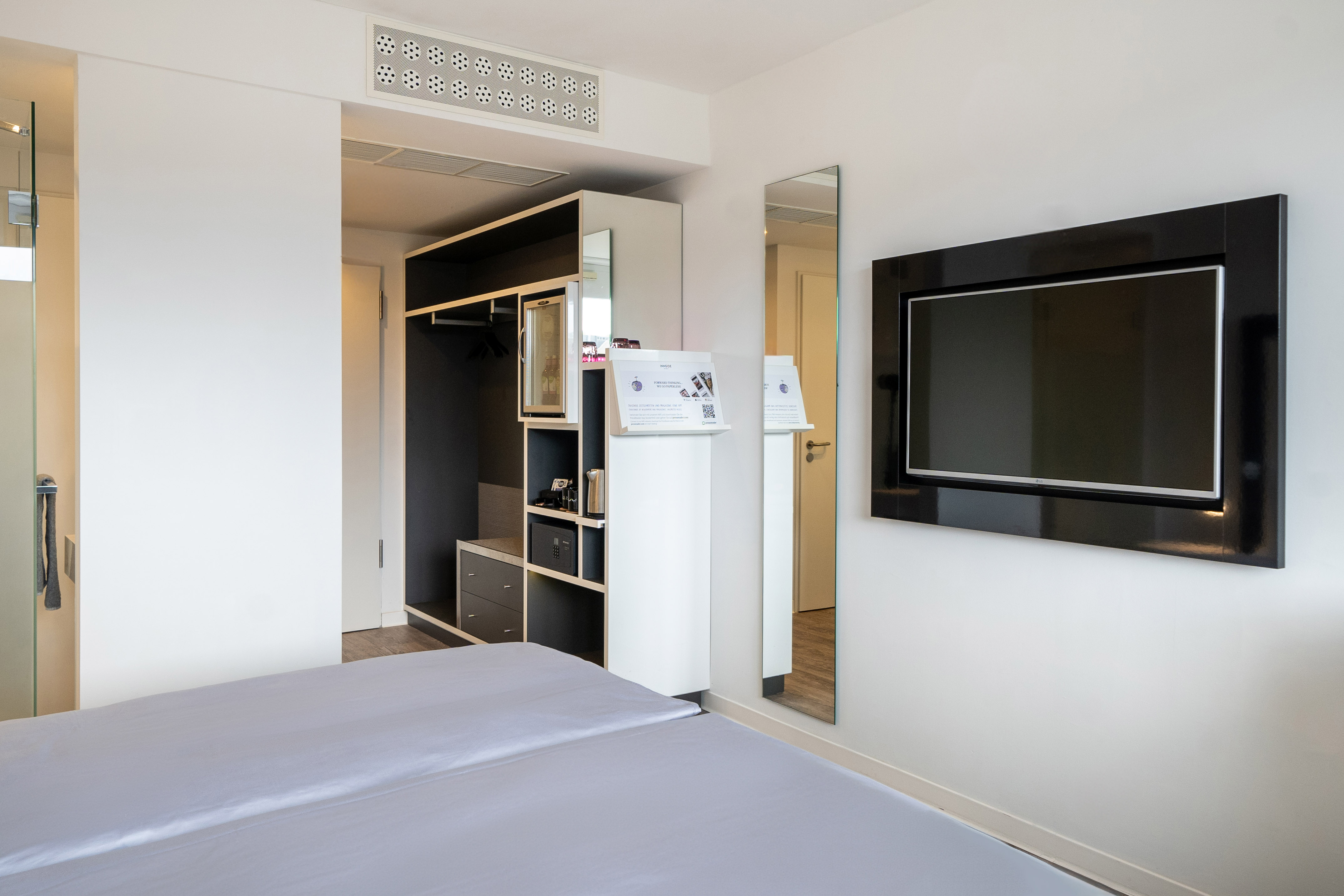 Modern hotel room: bed, wall-mounted TV, open wardrobe, minibar, mirror.