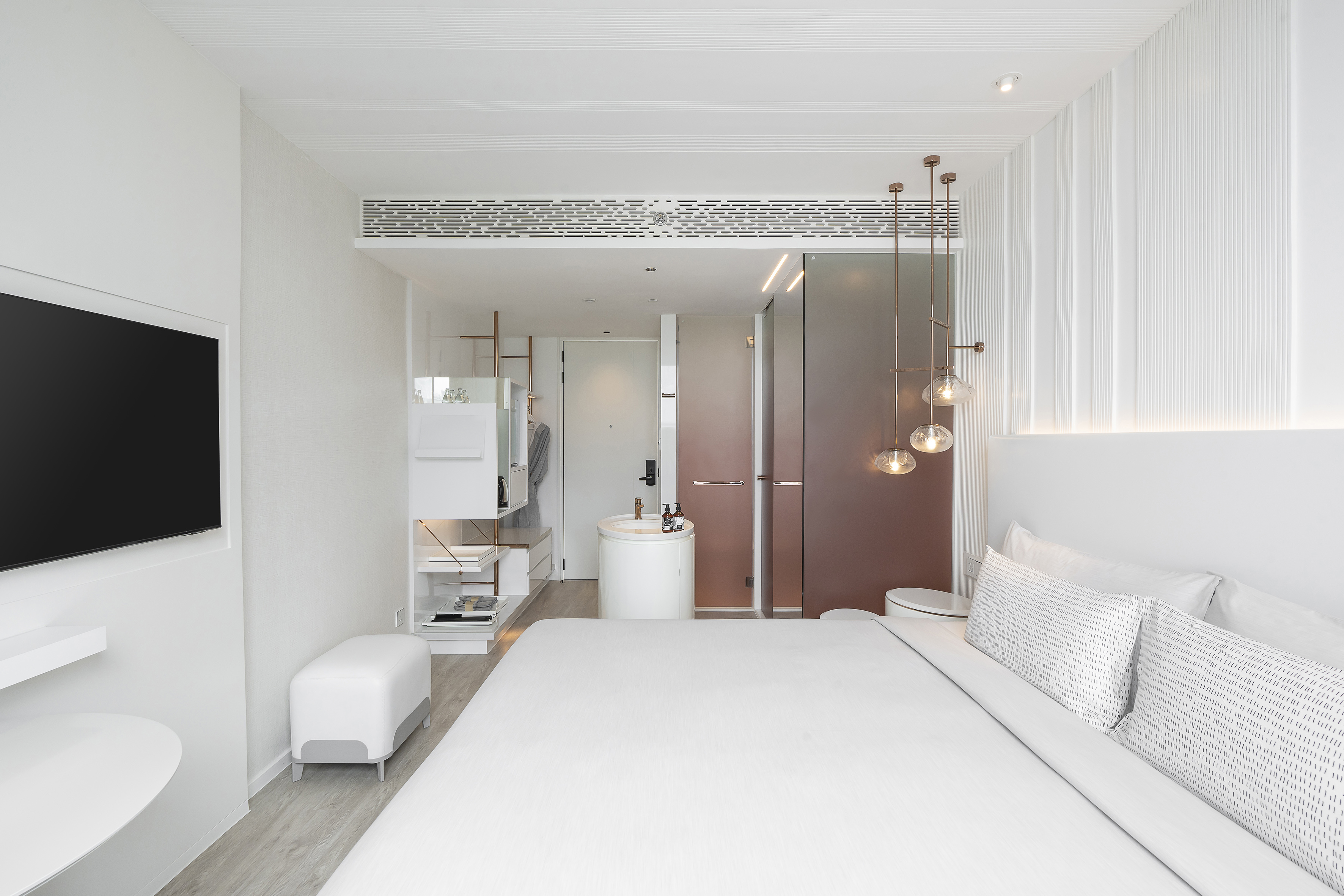 White minimalist hotel room with modern bed, TV, and copper pendant lights.