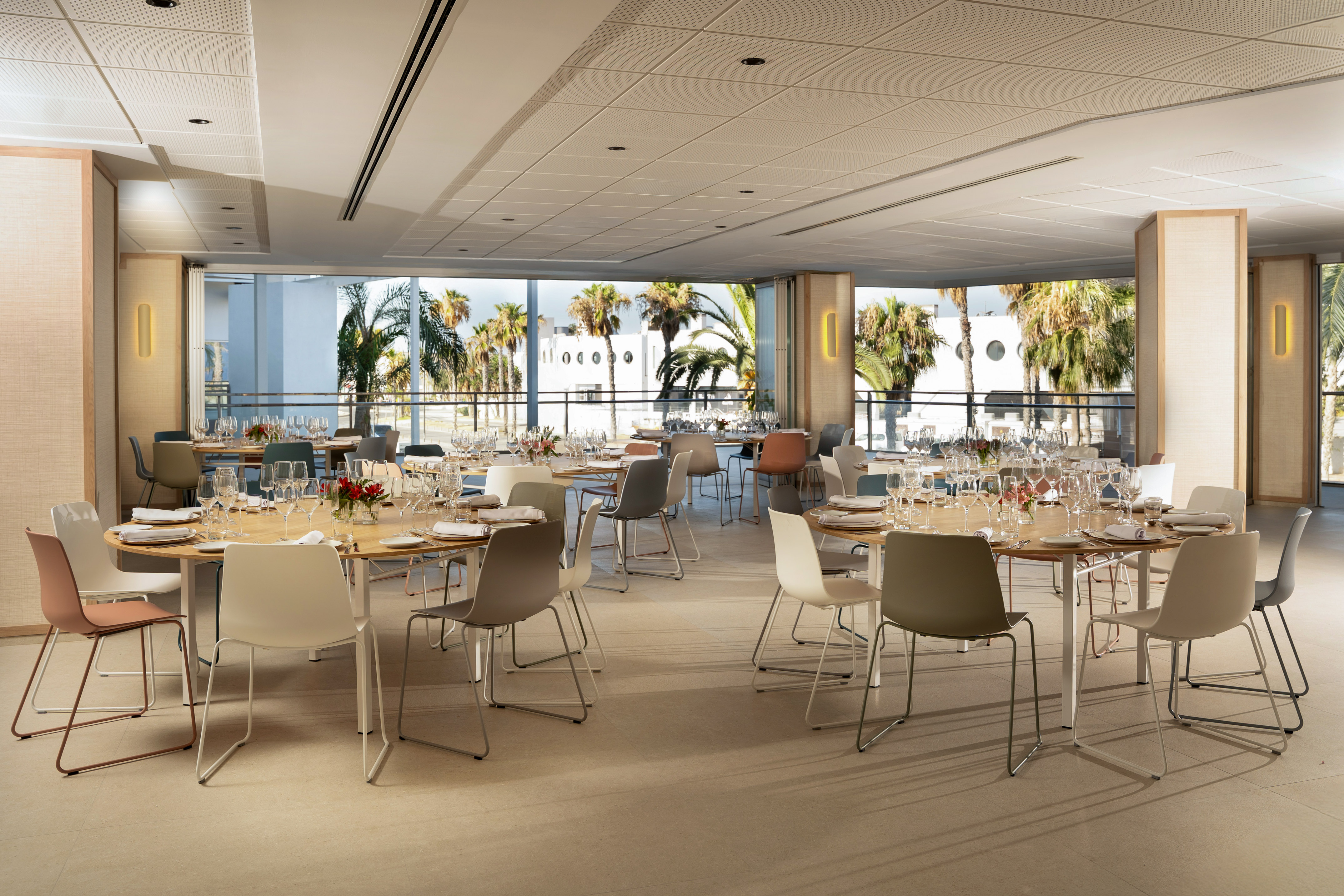 Modern restaurant dining room with round tables, colorful chairs, and palm tree view.