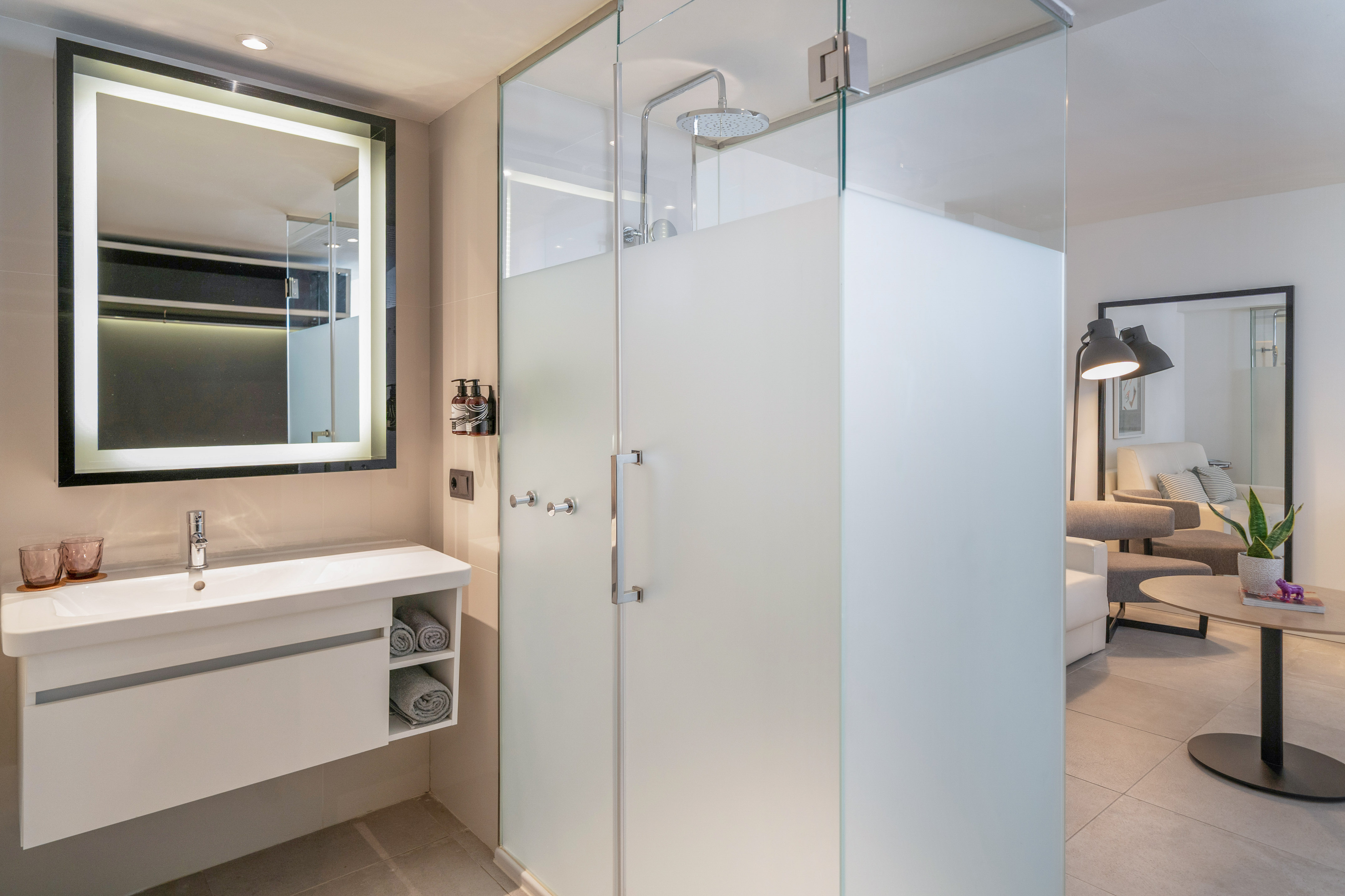 Modern hotel bathroom with illuminated mirror, vanity, and frosted glass shower.