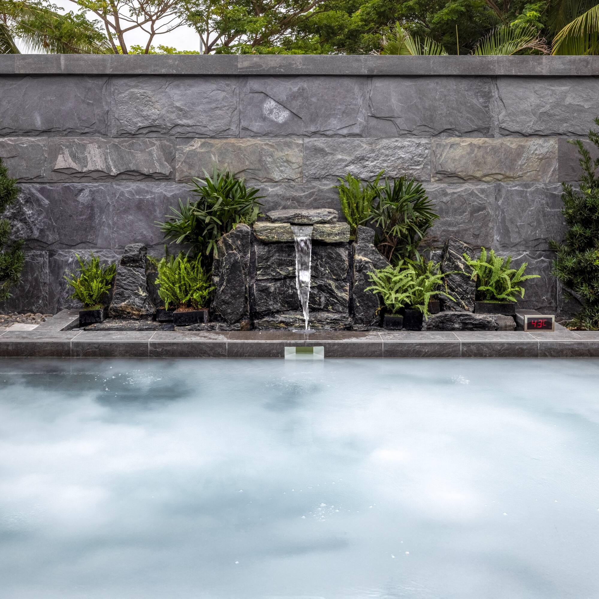Onsen hot tub with stone waterfall, lush plants, bamboo wall.