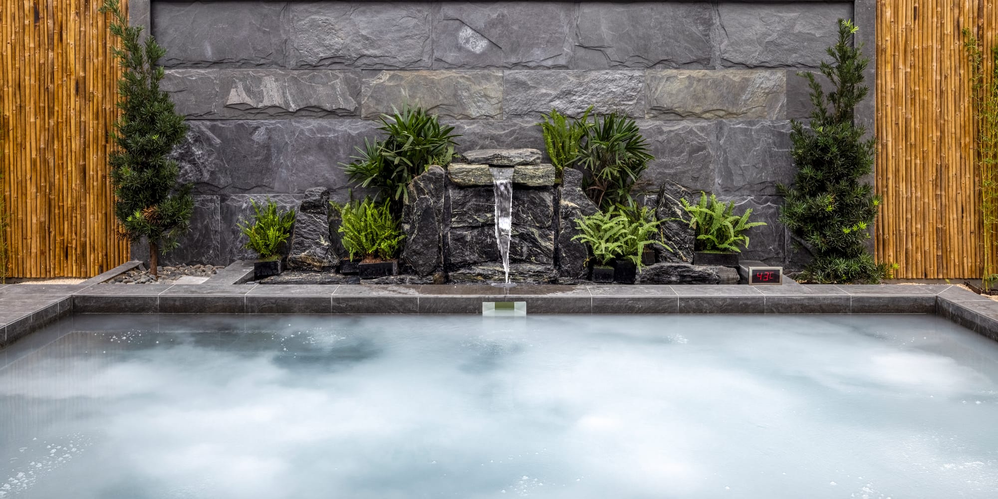 Outdoor hot tub with rock waterfall, lush plants, and bamboo wall.