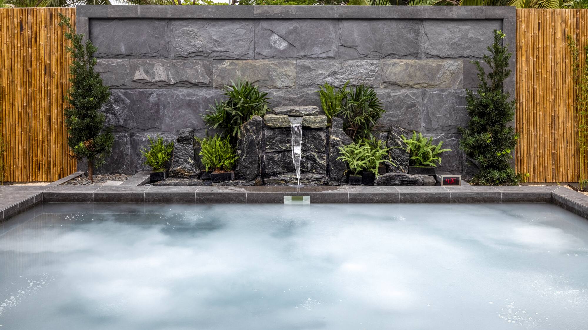 Outdoor hot tub with rock waterfall, lush plants, and bamboo wall.
