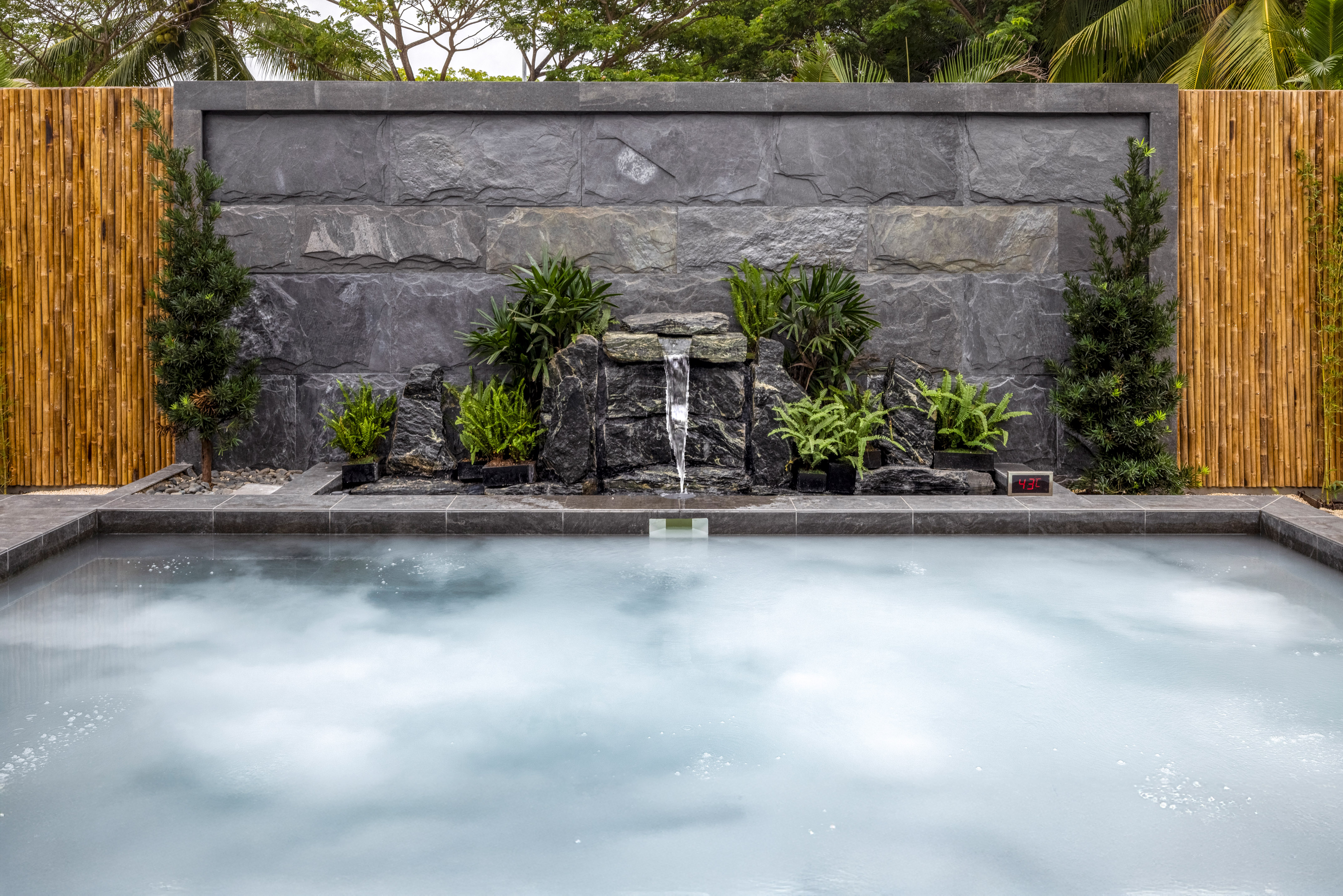 Onsen hot tub with stone waterfall, lush plants, bamboo wall.