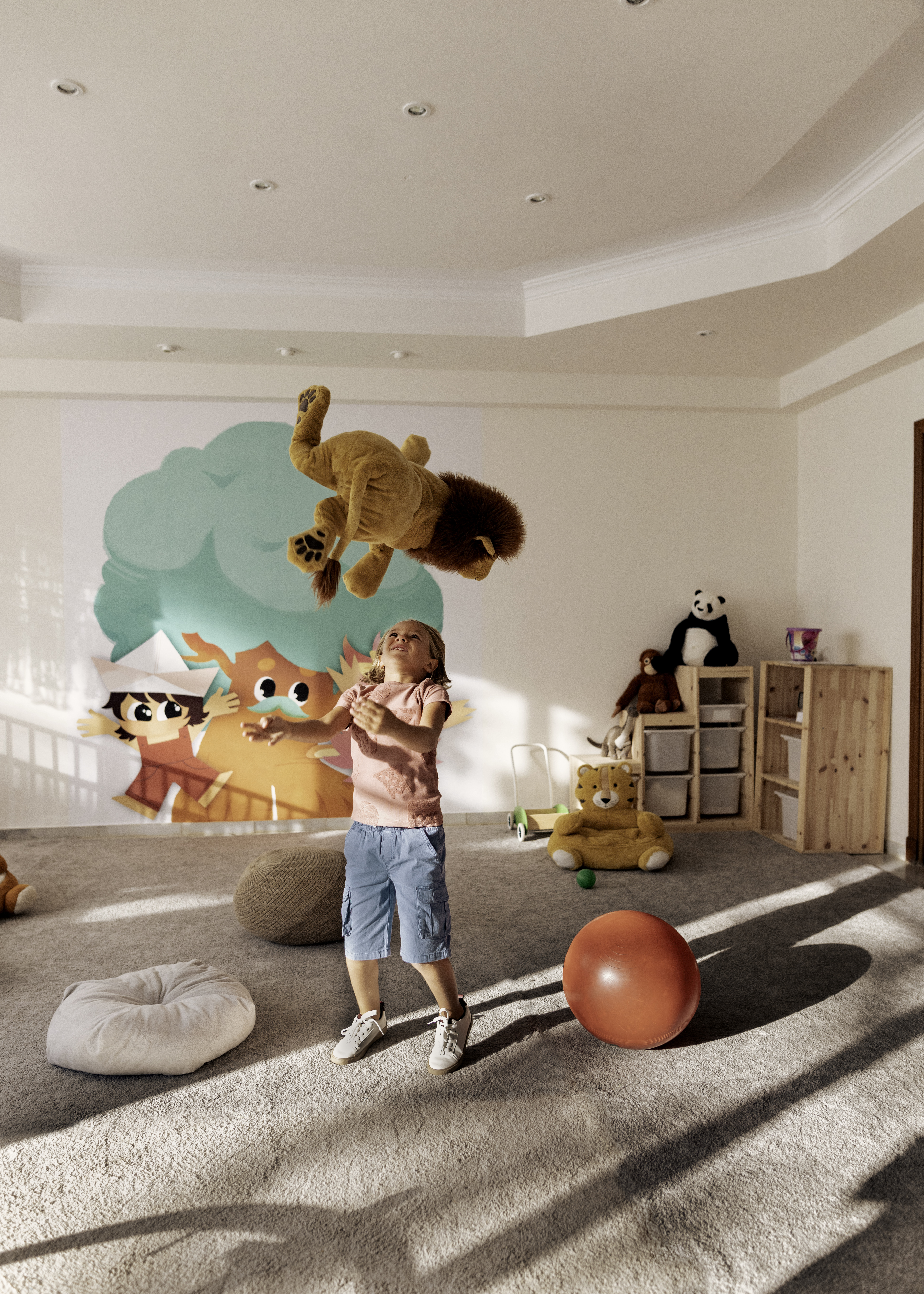 Child tossing lion plush toy in playroom with colorful character mural.
