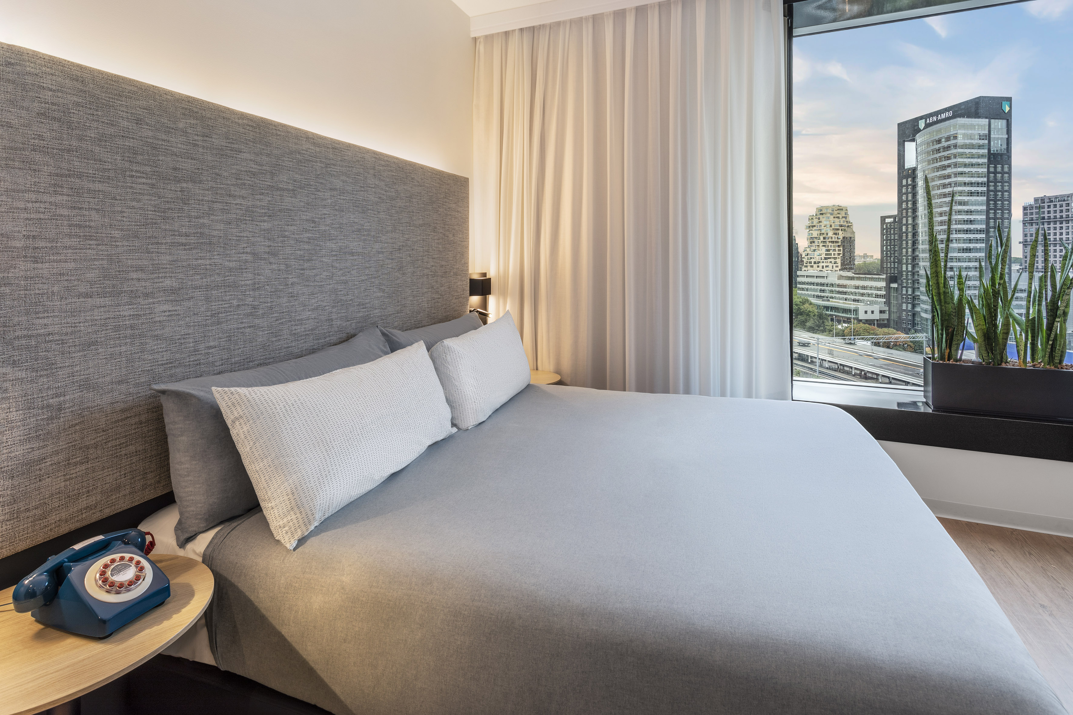 Modern hotel room with grey bed, city view window, and vintage blue phone.