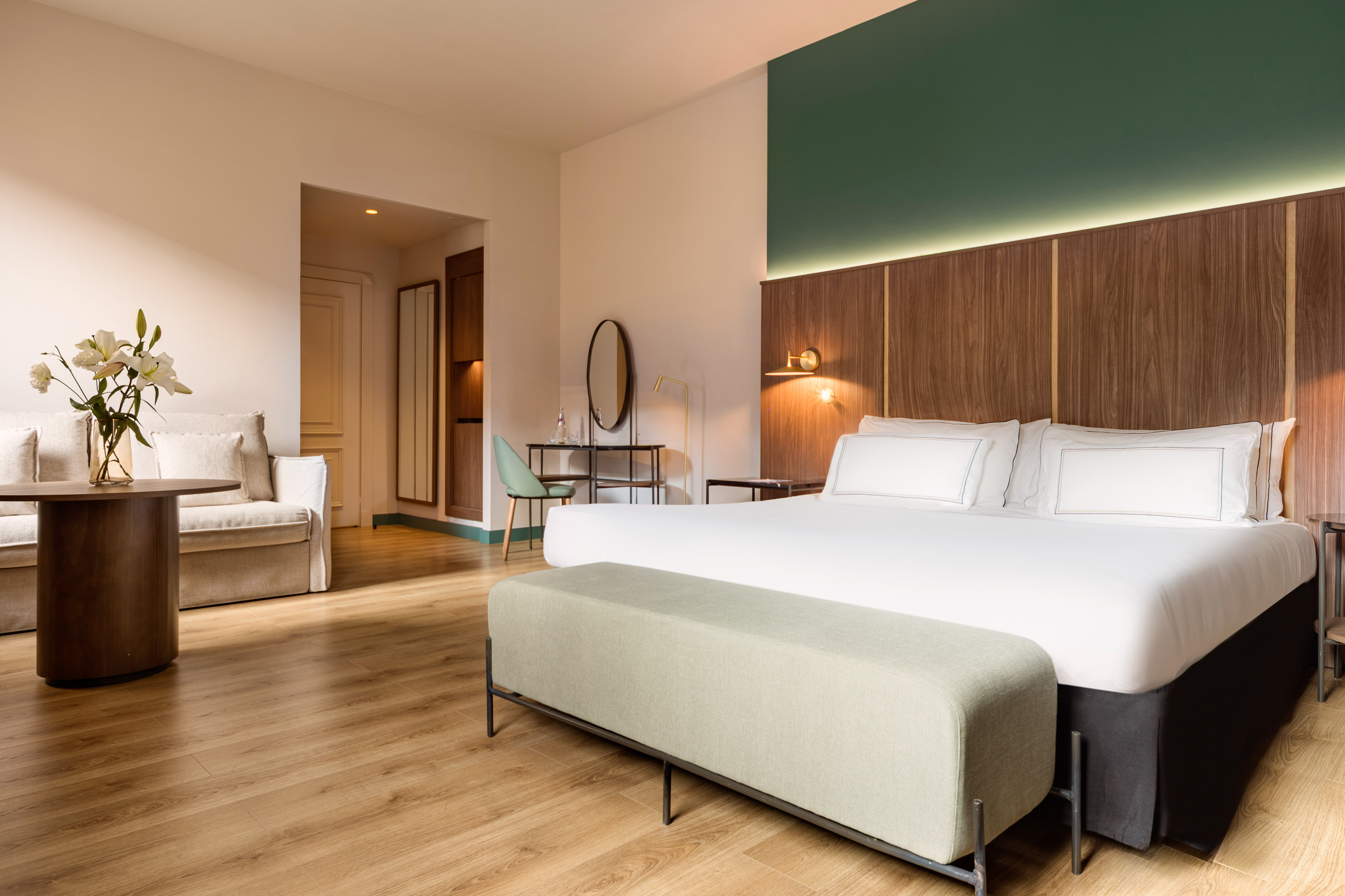 Modern hotel bedroom with king bed, wooden headboard, green wall, and lounge.