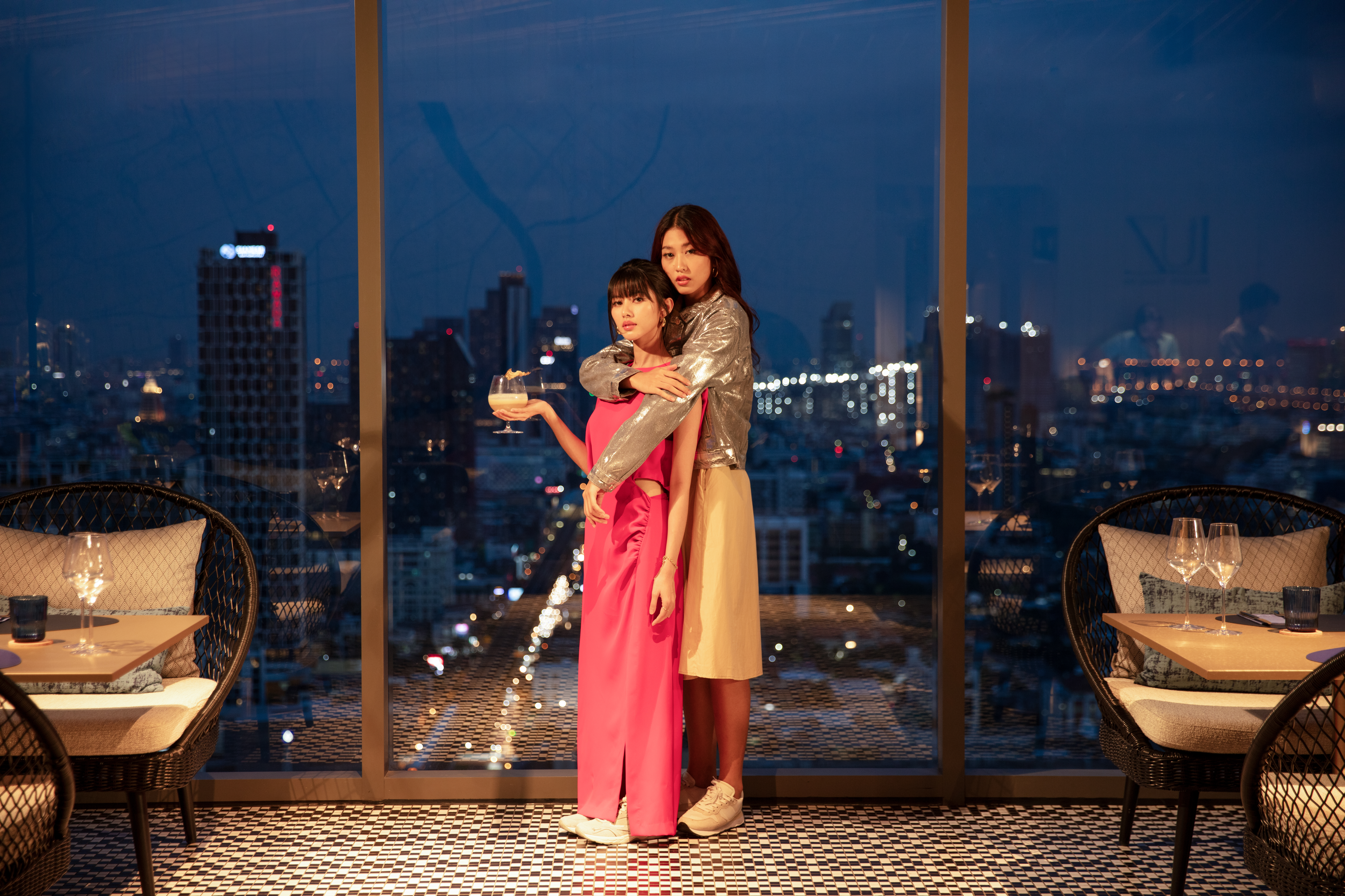 Two women embracing at modern city rooftop bar with night skyline view.