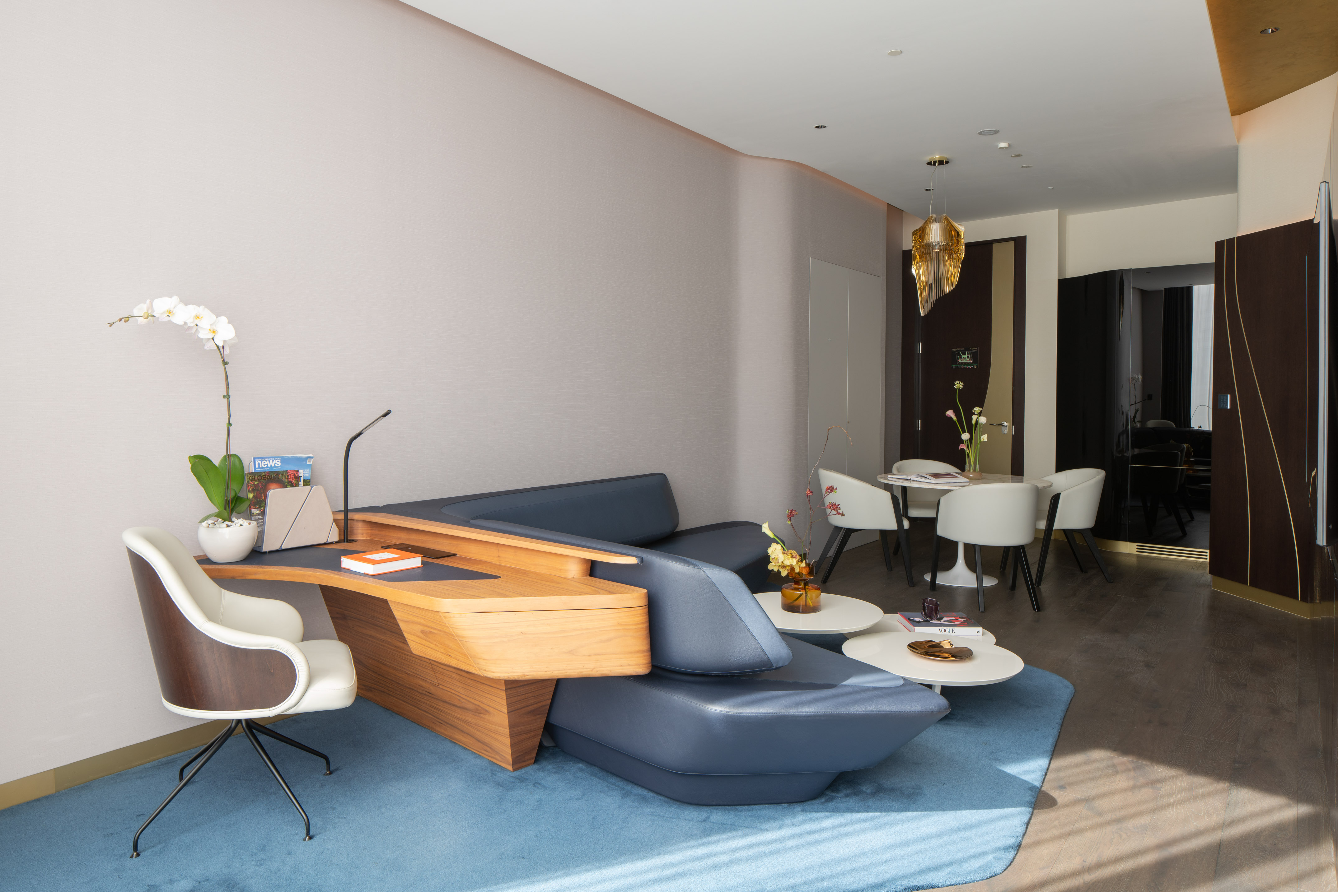 Modern living room with blue sofa, integrated wooden desk, white chair, and dining area.