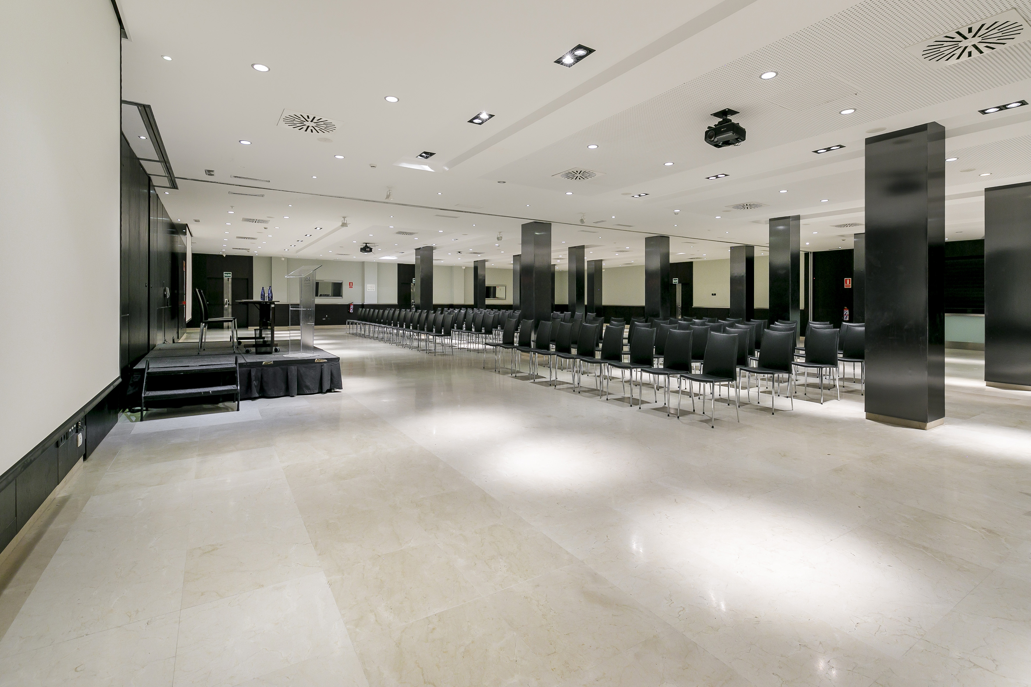 Empty conference room with rows of black chairs, stage, and projector screen.