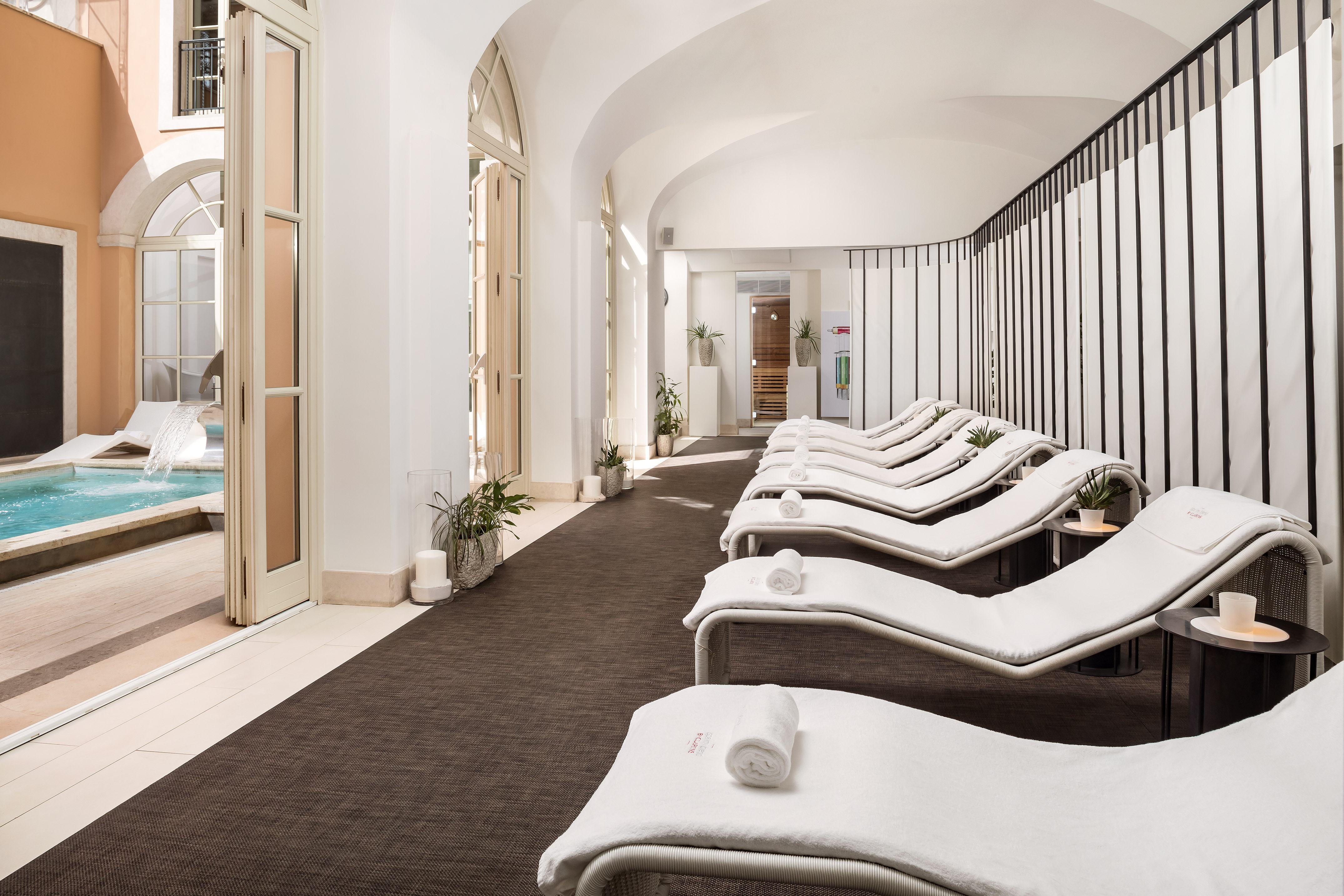 Luxury spa lounge with white chaises, outdoor pool view, and sauna.