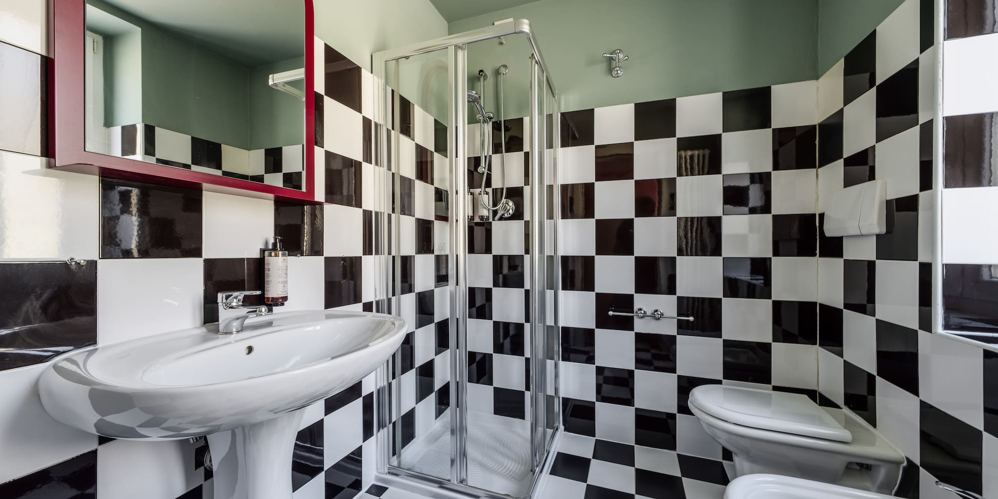 Black and white checkered bathroom with sink, shower, toilet, bidet, red mirror.