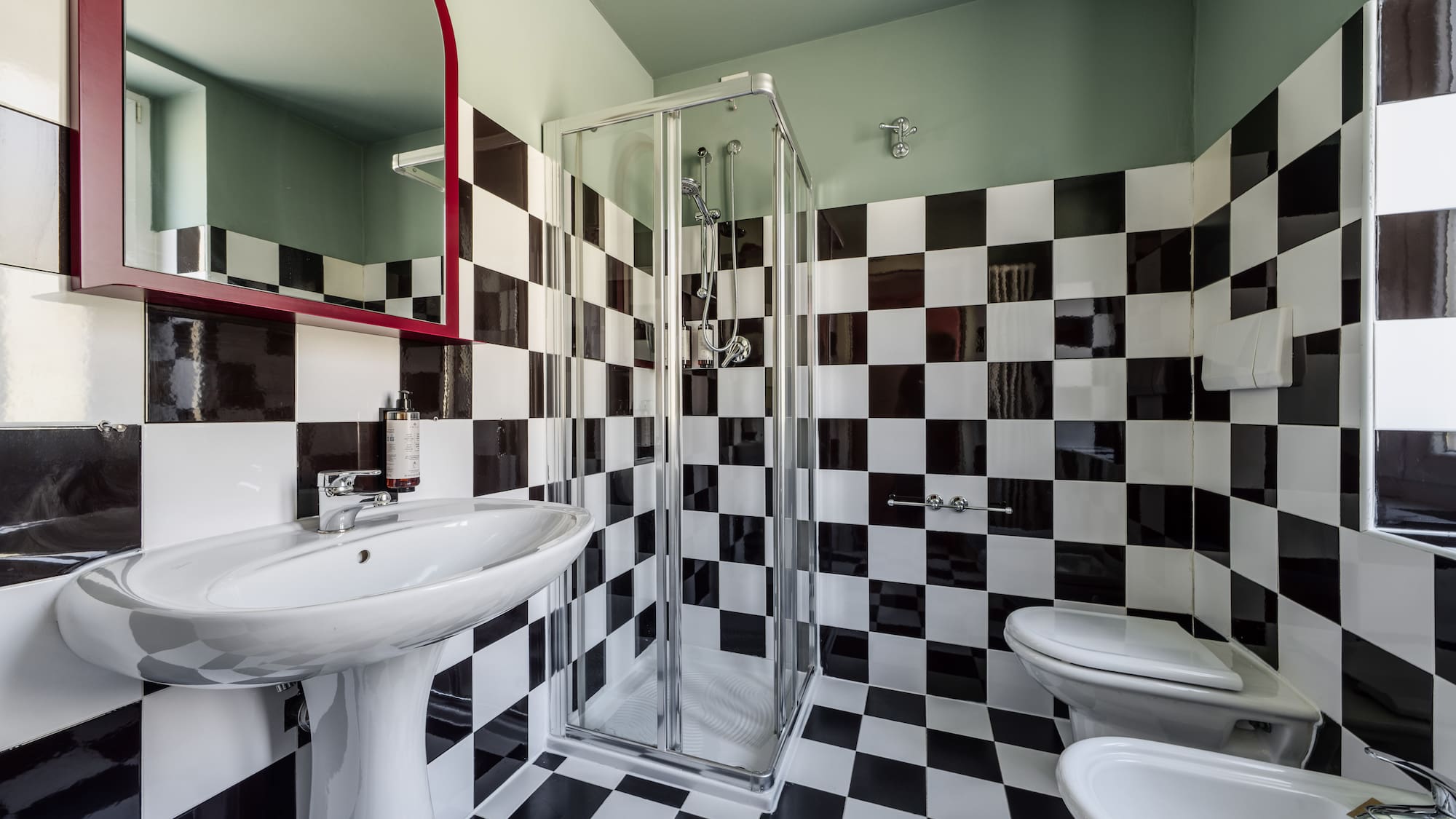 Black & white checkered bathroom with sink, shower, toilet, bidet, red mirror.