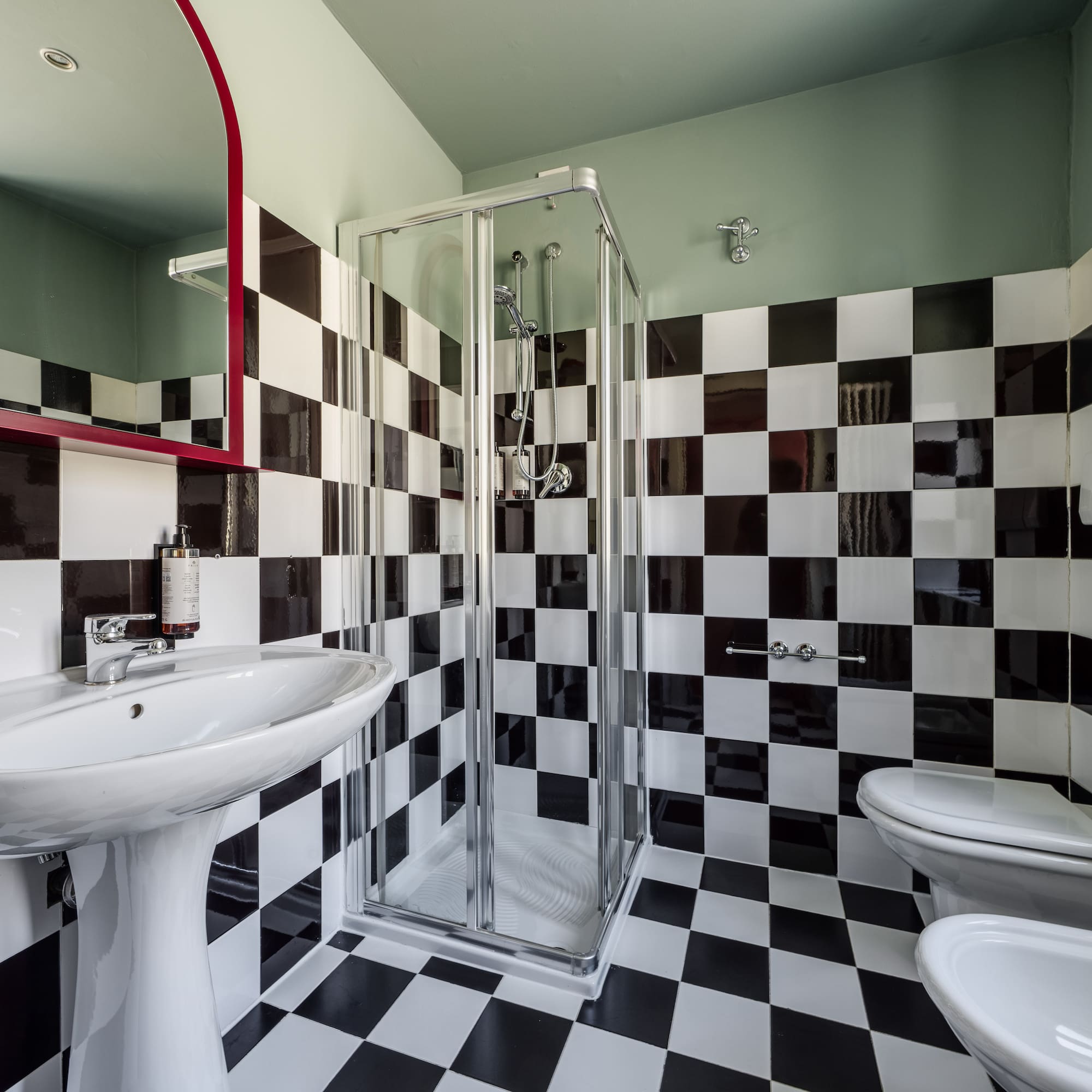 Black and white checkered bathroom with sink, shower, toilet, bidet, red mirror.