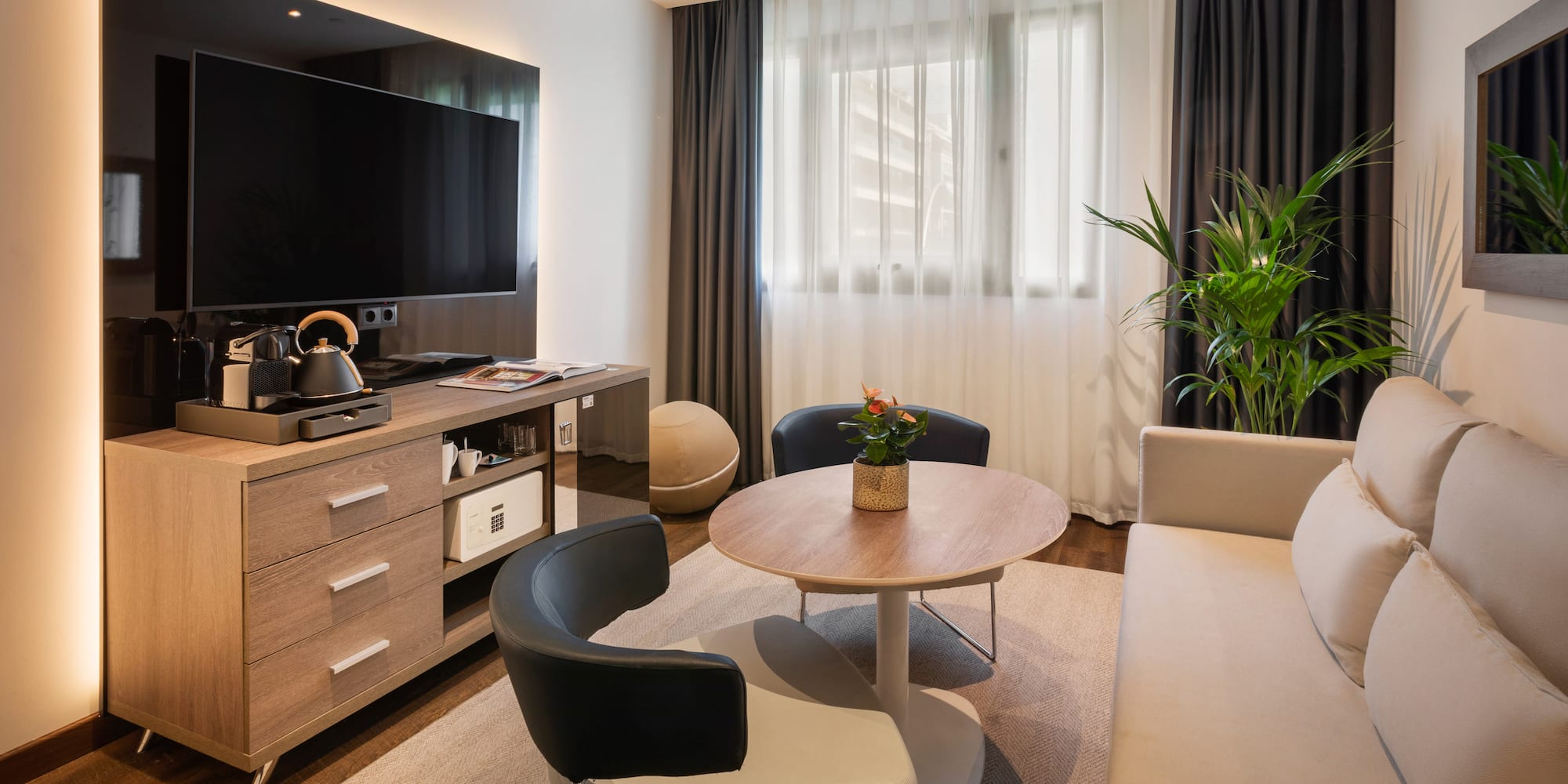 Moderna sala de estar de hotel con TV, sofá, sillones, mesa y cafetera.