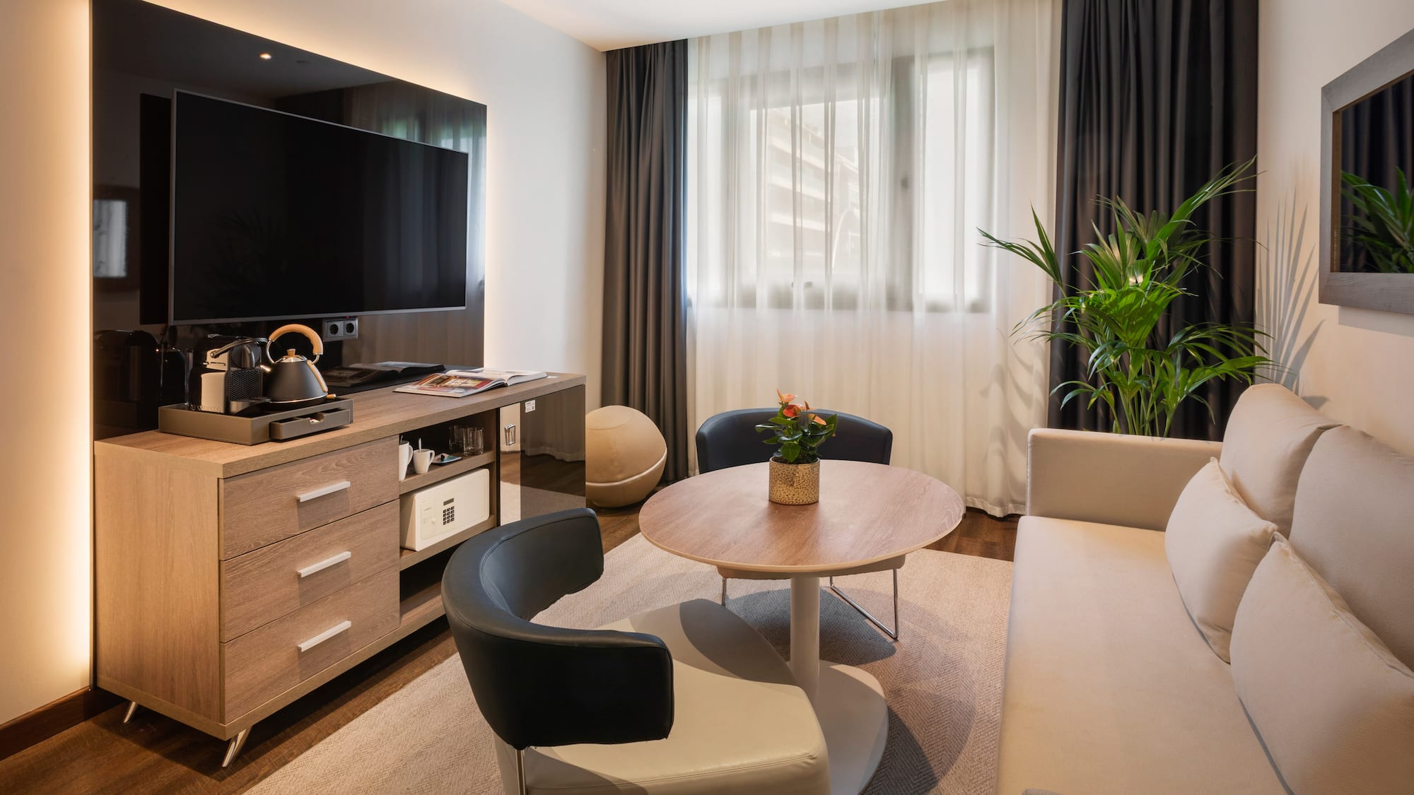 Moderna sala de estar de hotel con TV, sofá, sillones, mesa y cafetera.