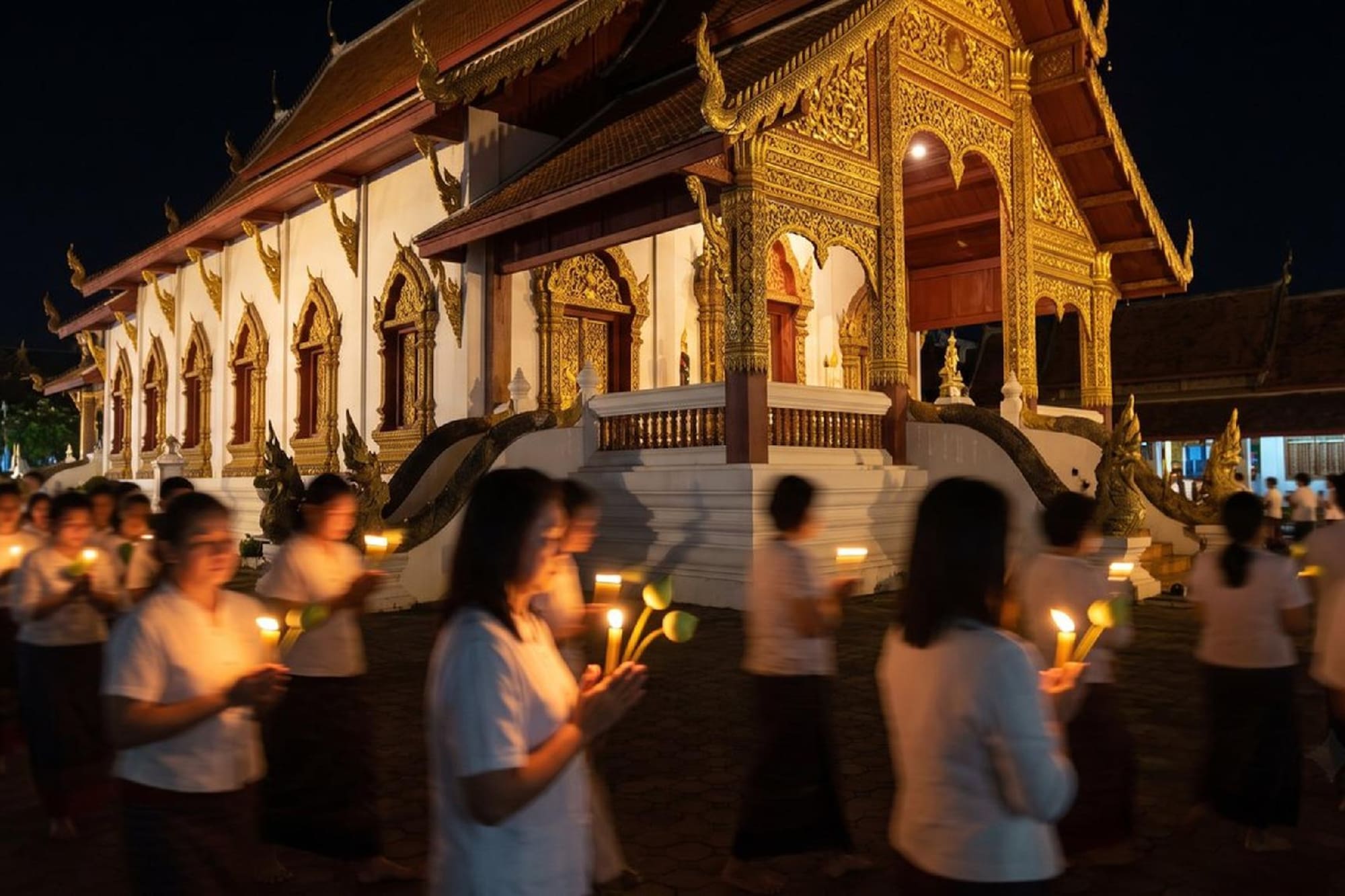 Thai temple night ceremony with people holding candles and lotus flowers.