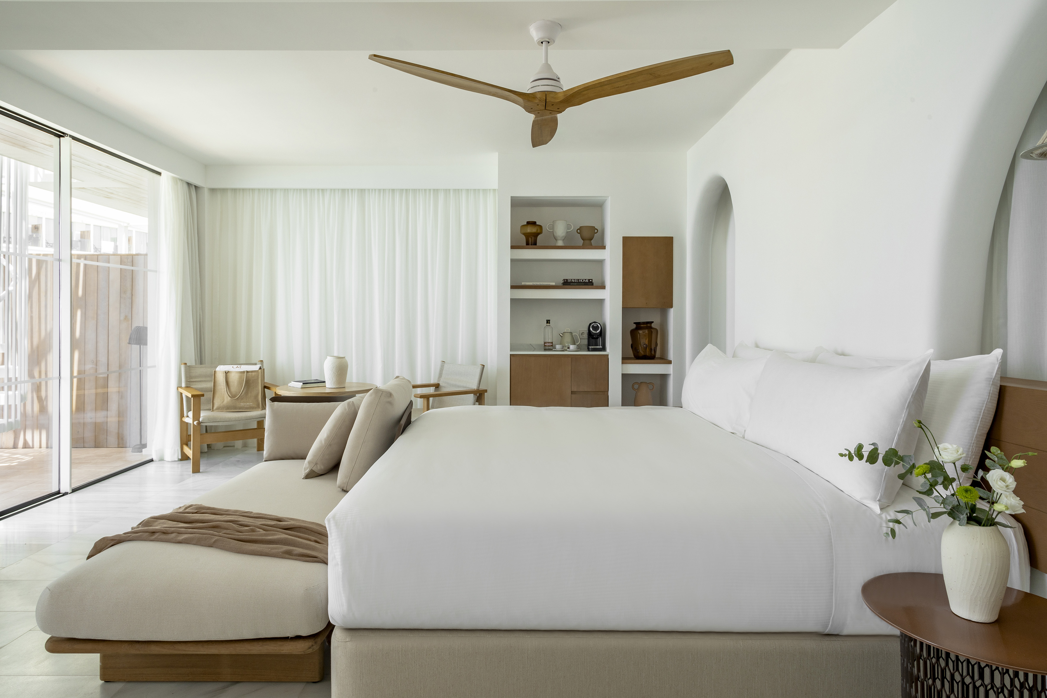 Minimalist hotel bedroom with white bed, wooden fan, and large patio doors.