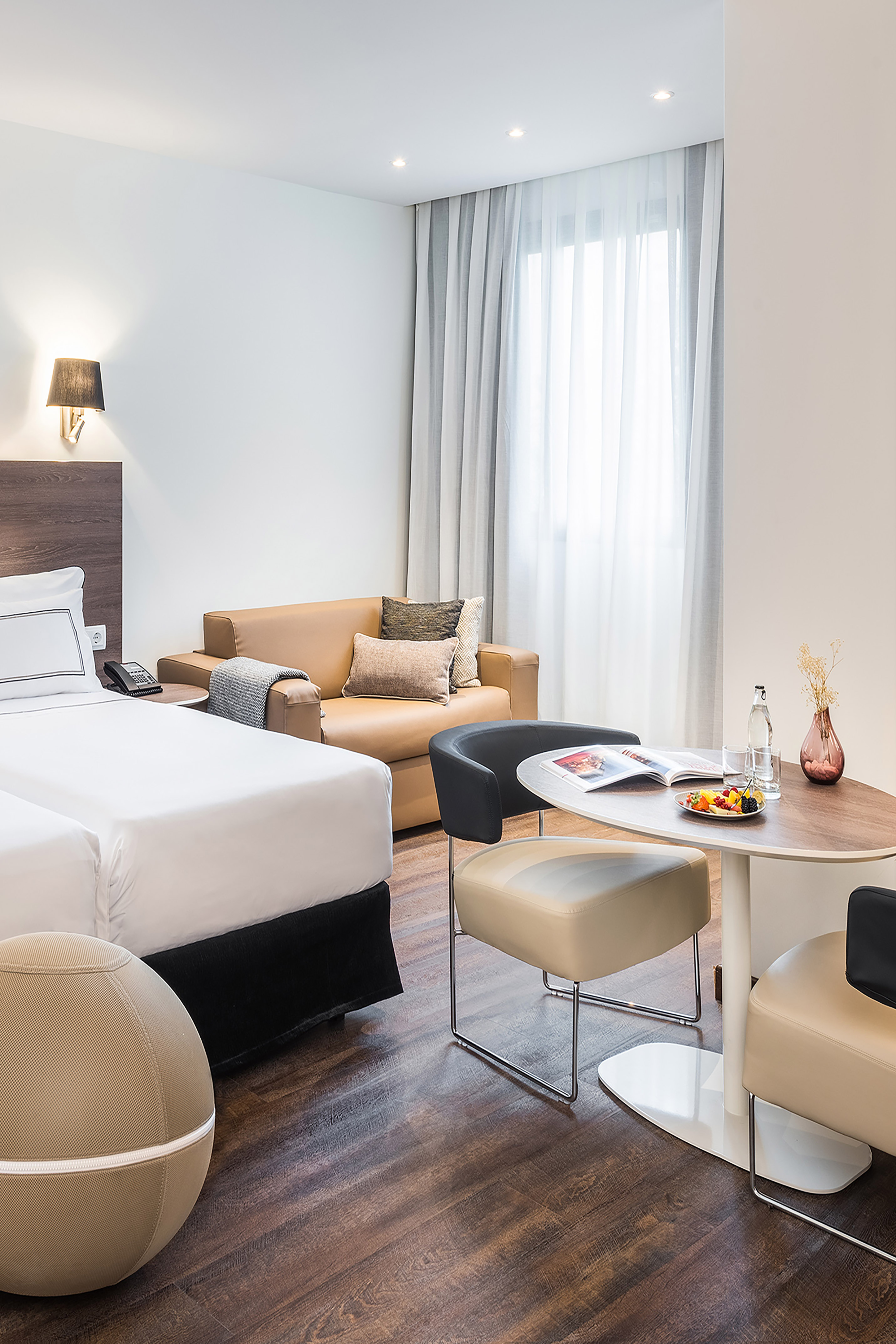 Modern hotel suite with white bed, tan sofa, dining table, and fresh fruit.