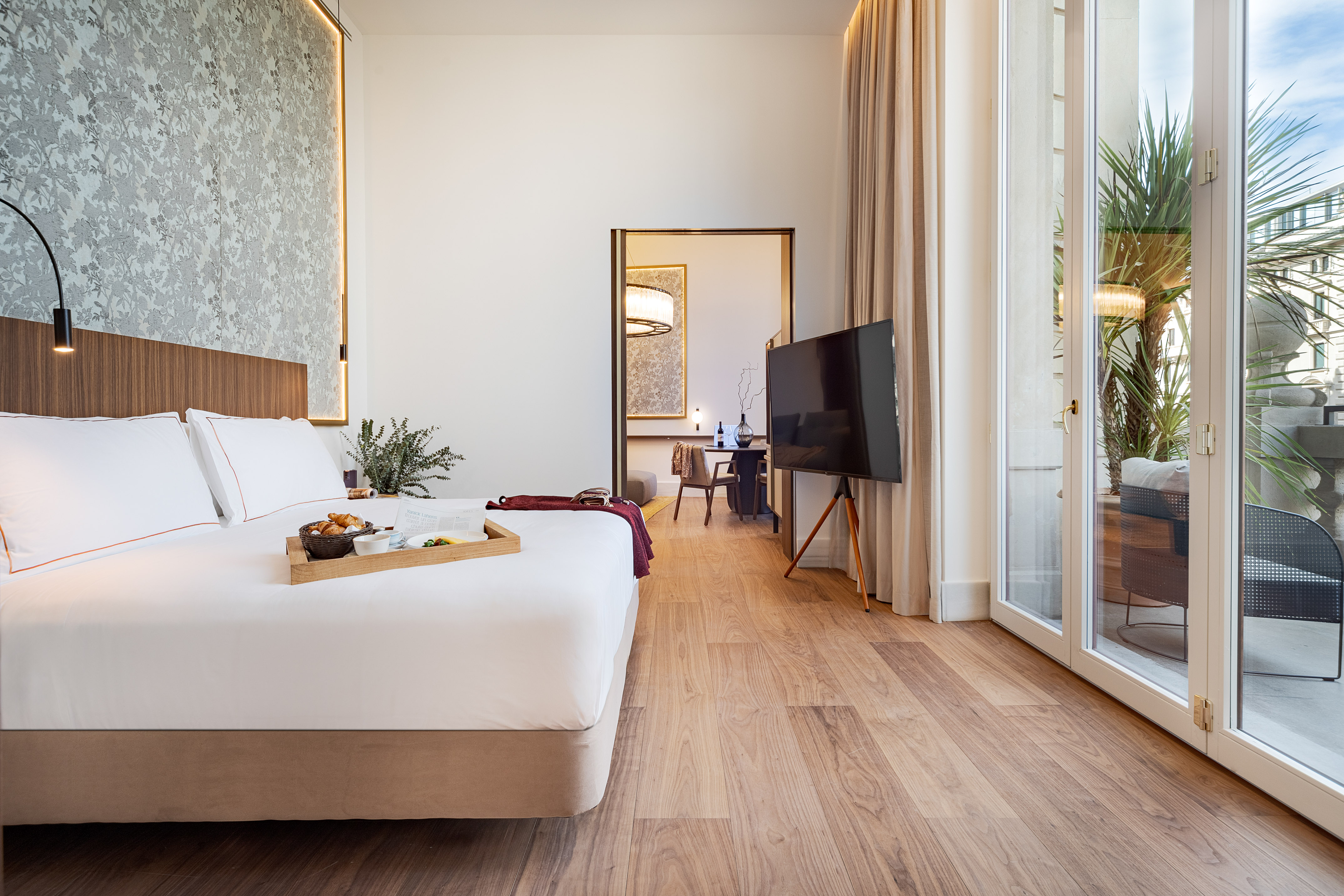 Modern hotel suite with bed, breakfast tray, TV, wood floors, and balcony.